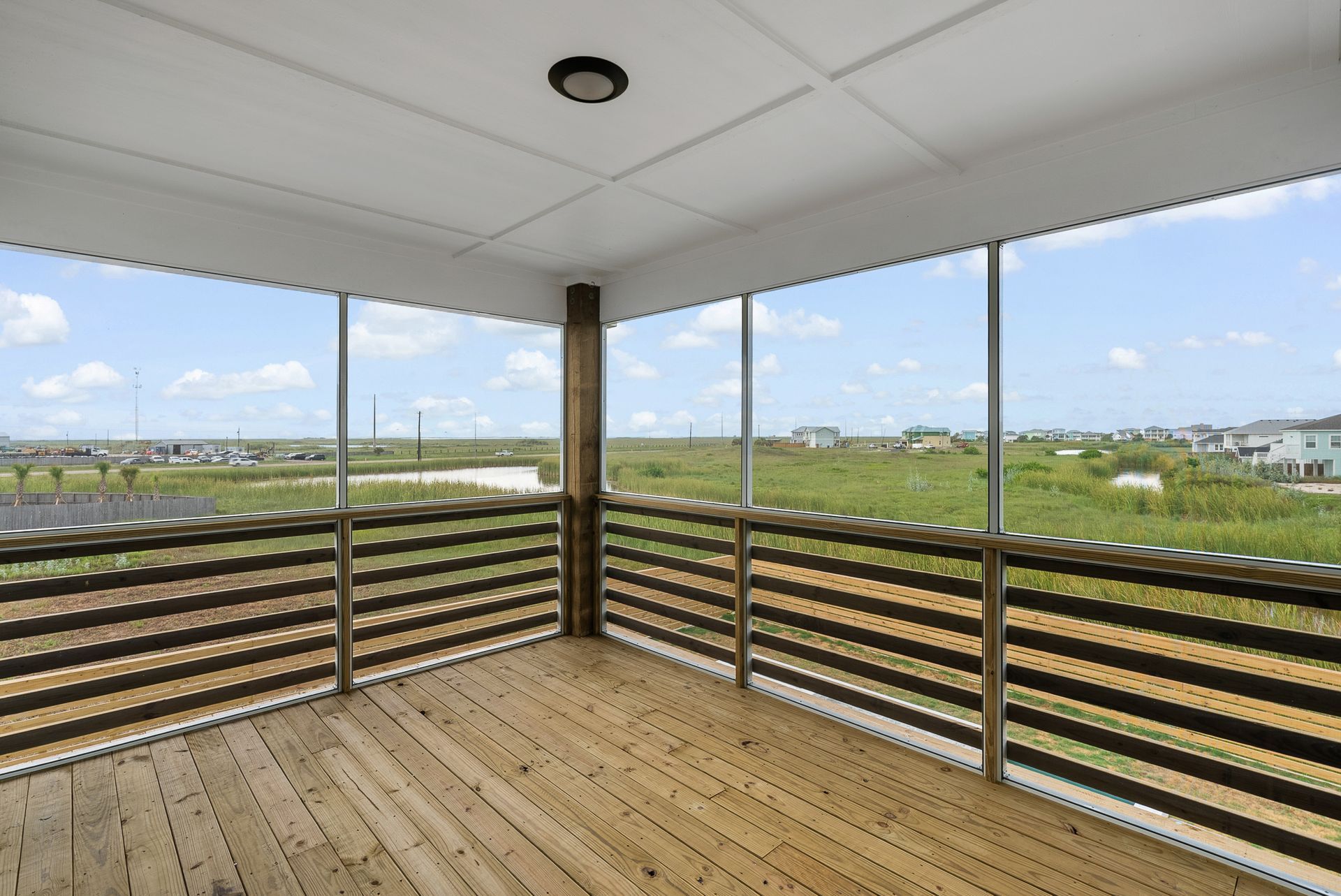 Wooden deck with railing and screened walls overlooking a grassy field and buildings under a cloudy sky.