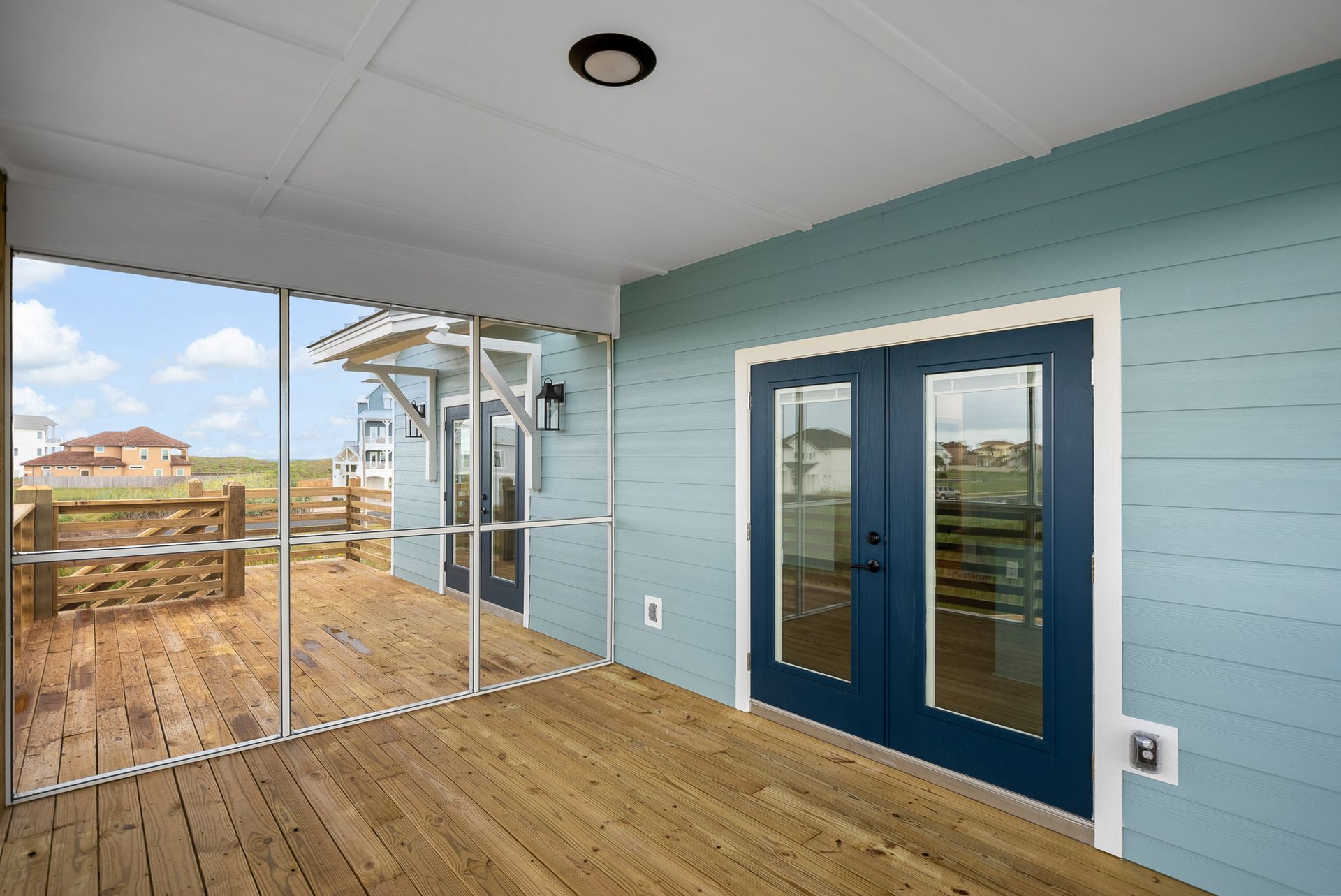 Screened porch with blue siding, wooden deck, and double doors; view of homes and sky.
