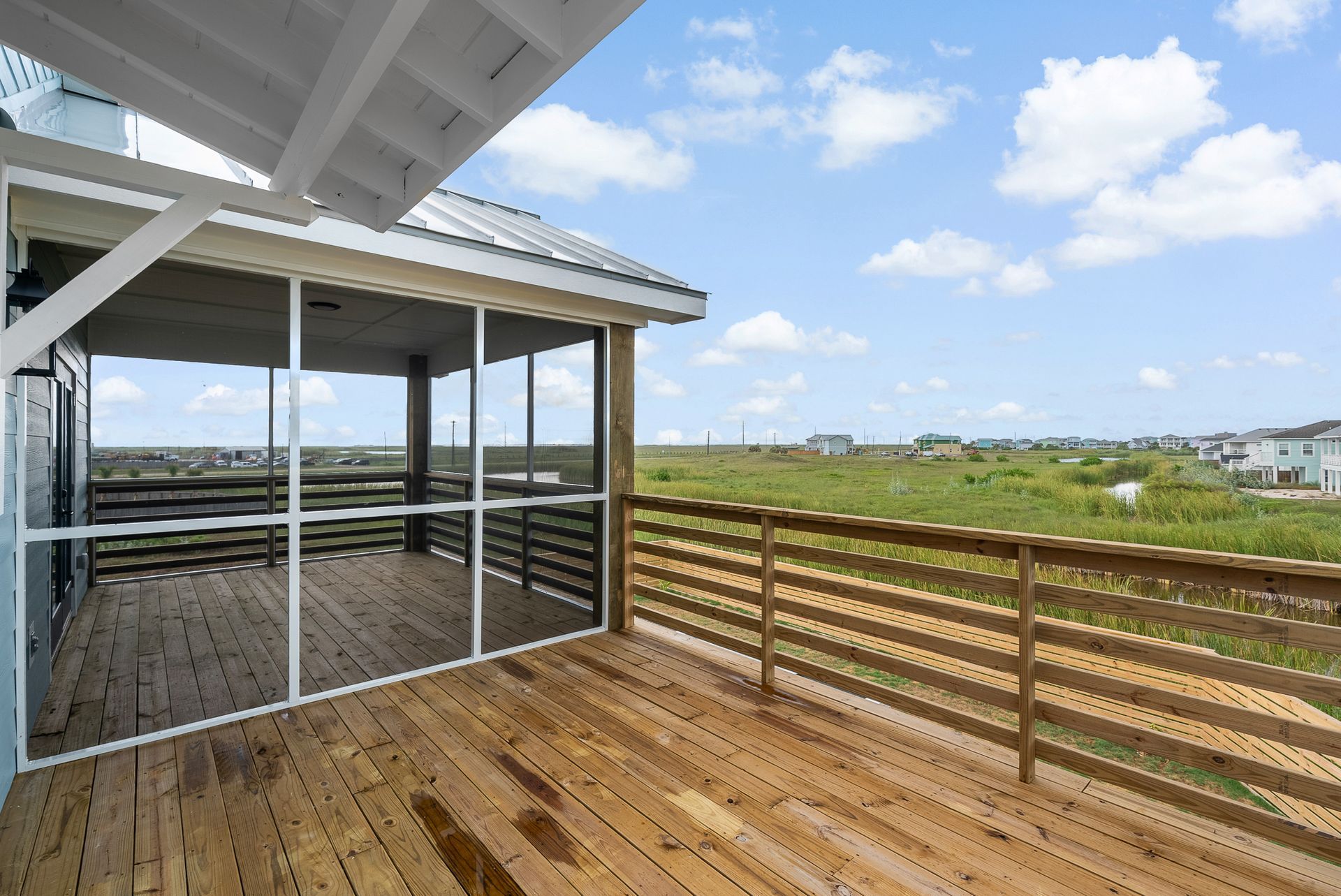 Wooden deck with screened porch, overlooking marsh and houses under a cloudy sky.
