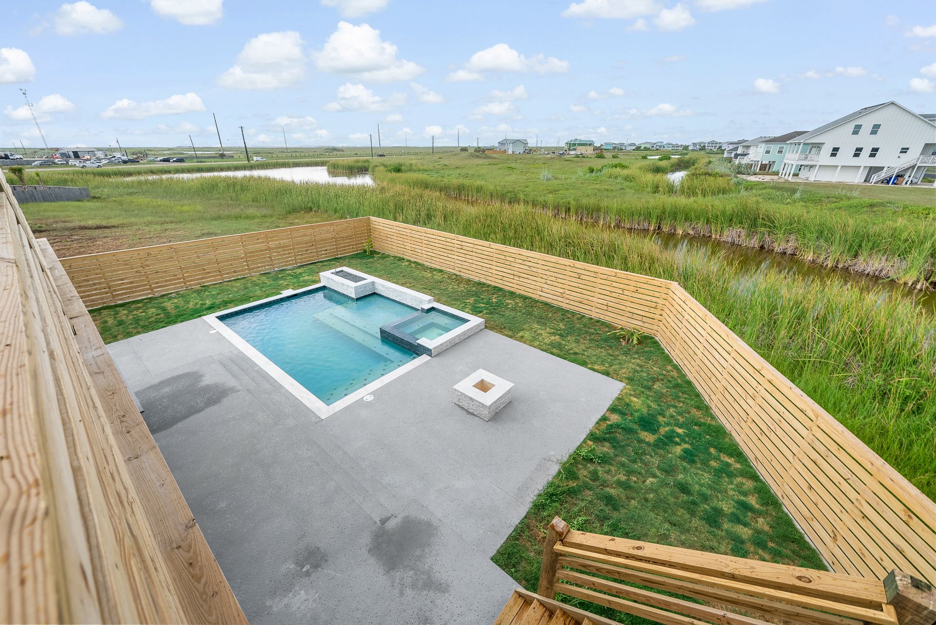 High-angle view of a backyard with a pool, hot tub, fire pit, and a wooden fence, surrounded by marshland.