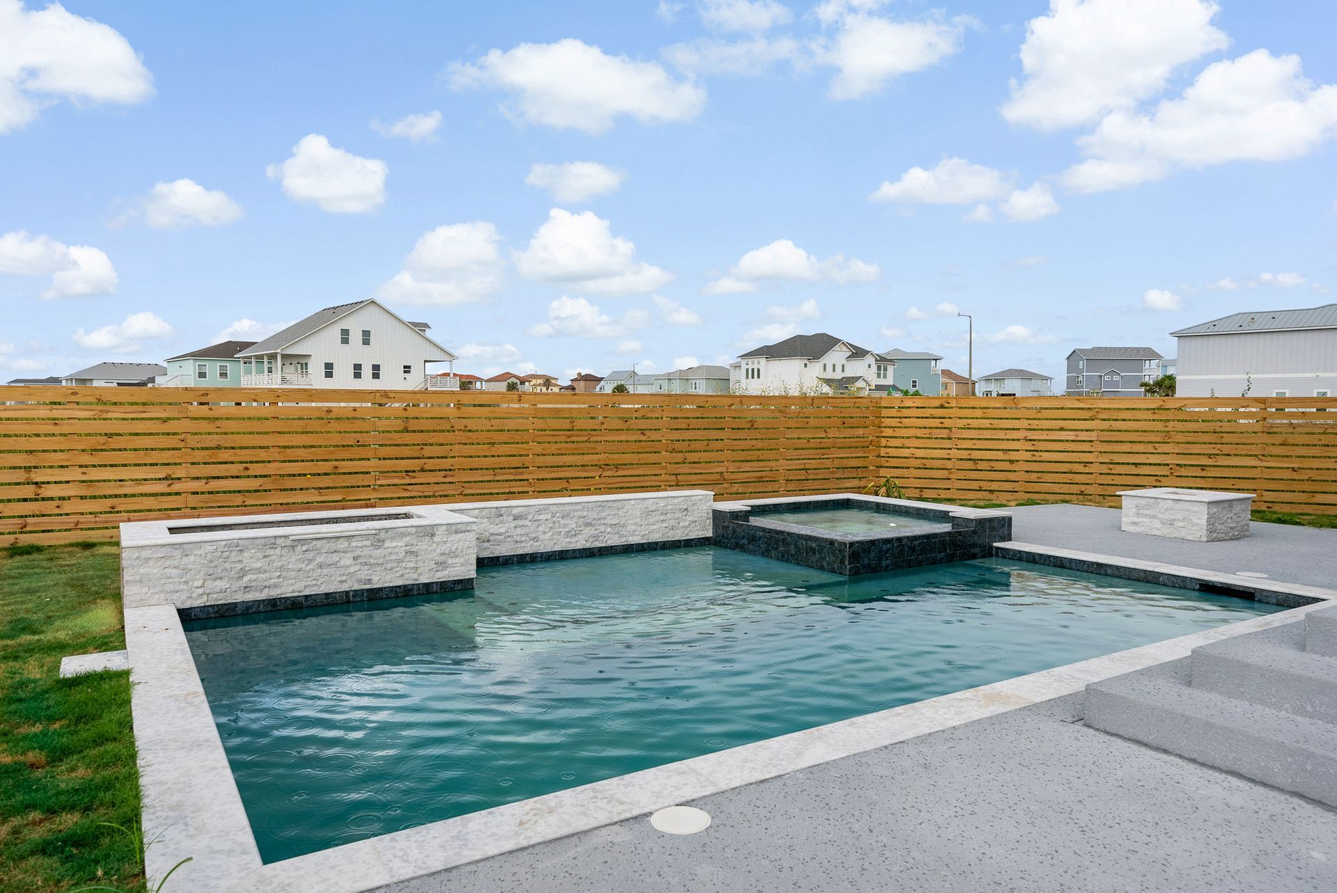 Backyard pool with spa, fire pit, and wooden fence against a sky with scattered clouds.
