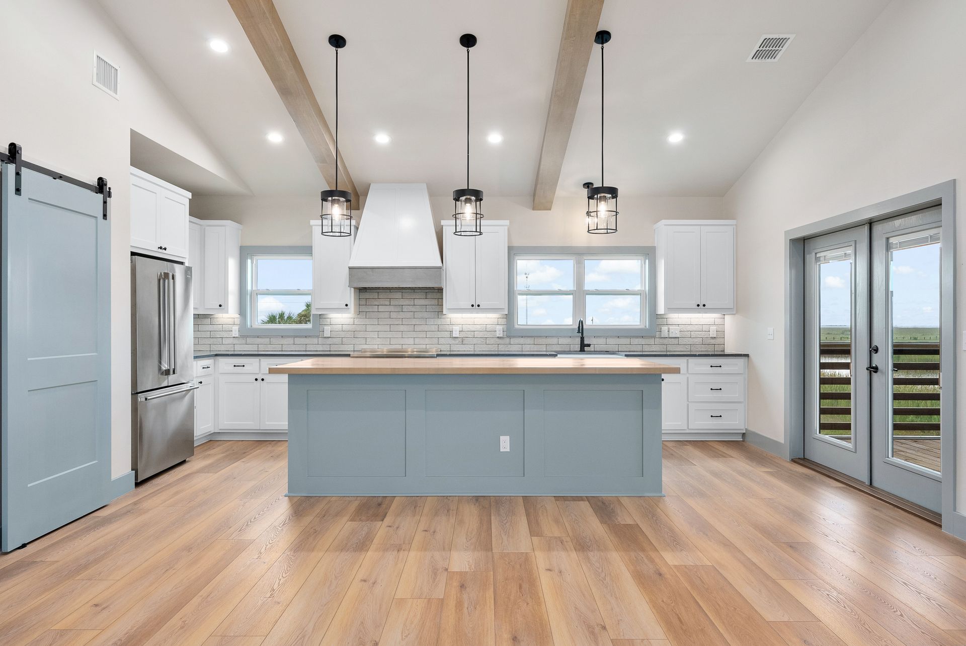 Spacious kitchen with blue island, white cabinets, wood beams, and large windows.