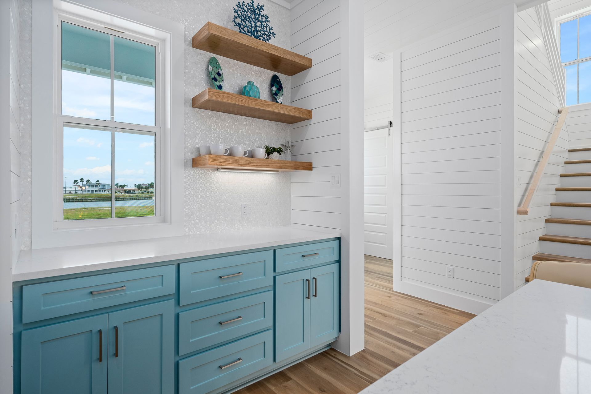 A kitchen with blue cabinets , white counter tops , wooden shelves and a window.