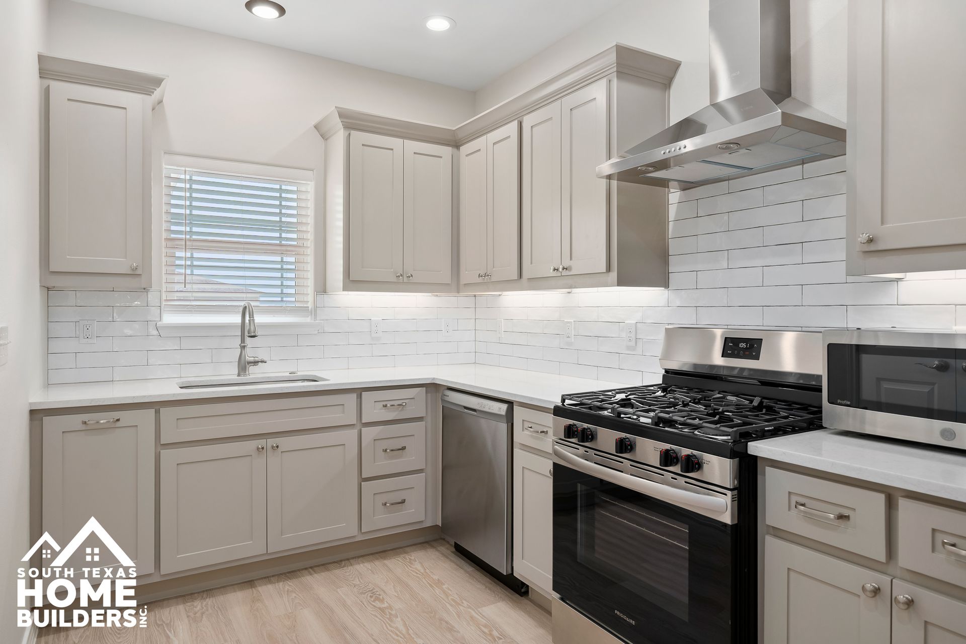 Modern kitchen with light gray cabinets, stainless steel appliances, and white brick-style backsplash.