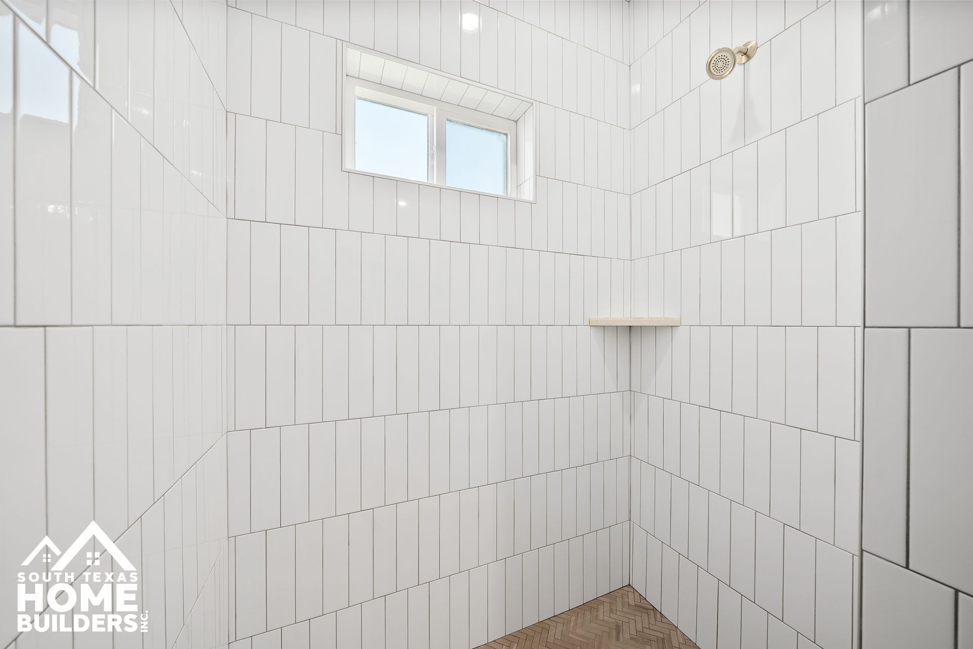 White tiled shower with small window, built-in shelf, and gold showerhead.