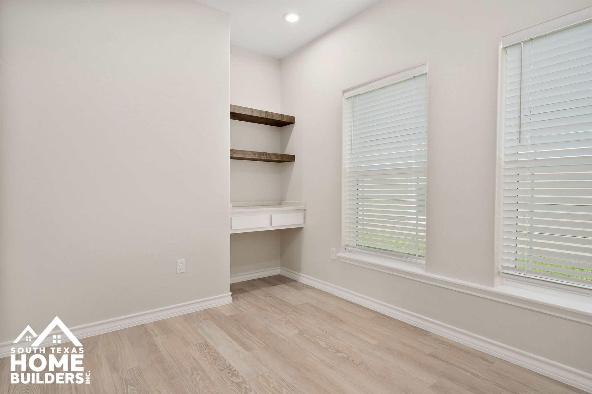 Empty room with a built-in desk, shelves, and two windows with blinds. Light wood flooring and neutral walls.