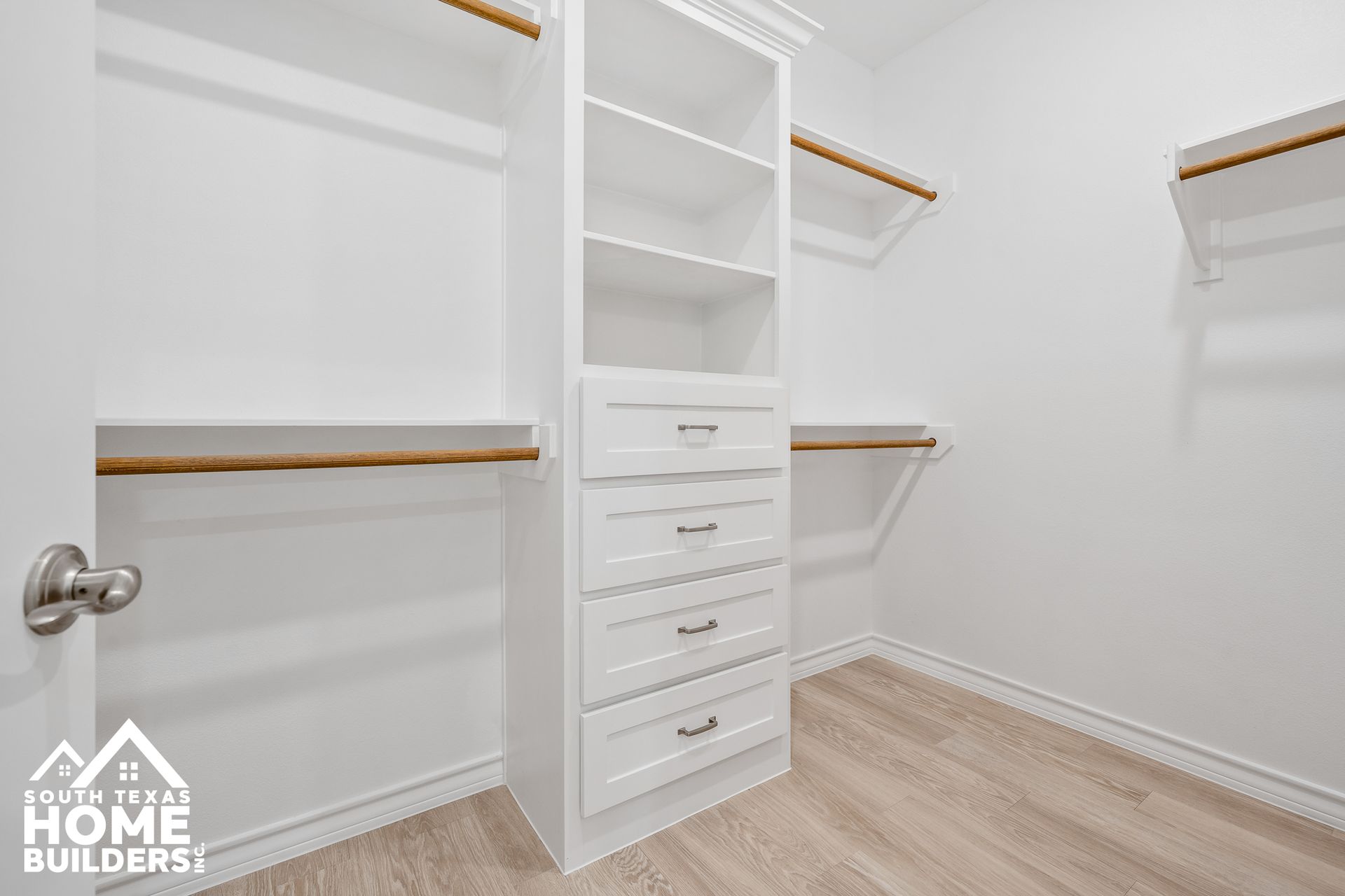 Walk-in closet with white shelves and drawers, wooden rods, and light flooring.