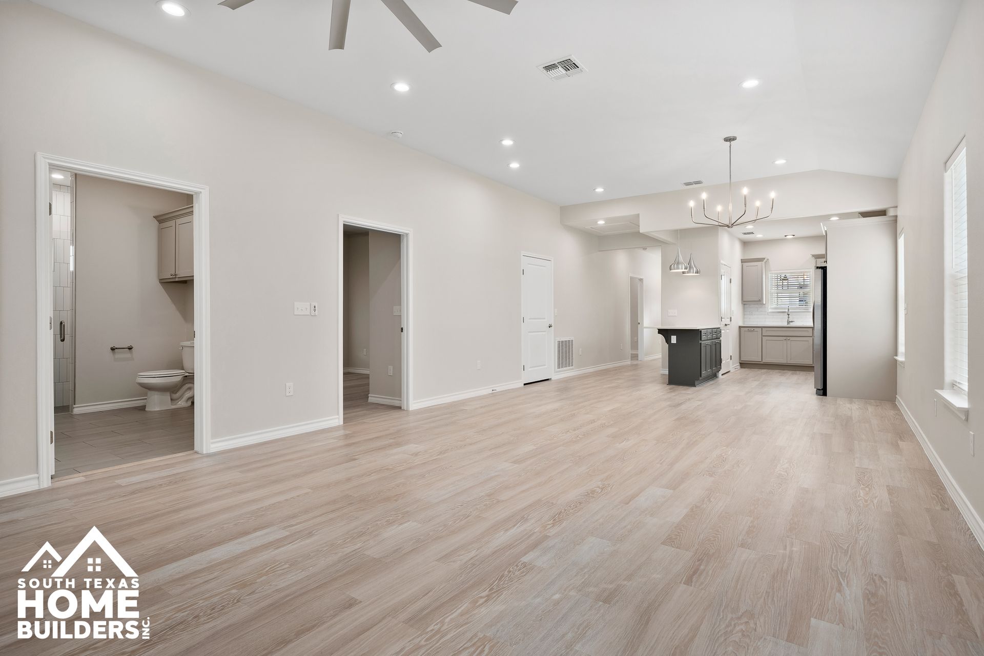Empty, renovated living room with light wood floors, recessed lighting, and a view of the kitchen and bathroom.