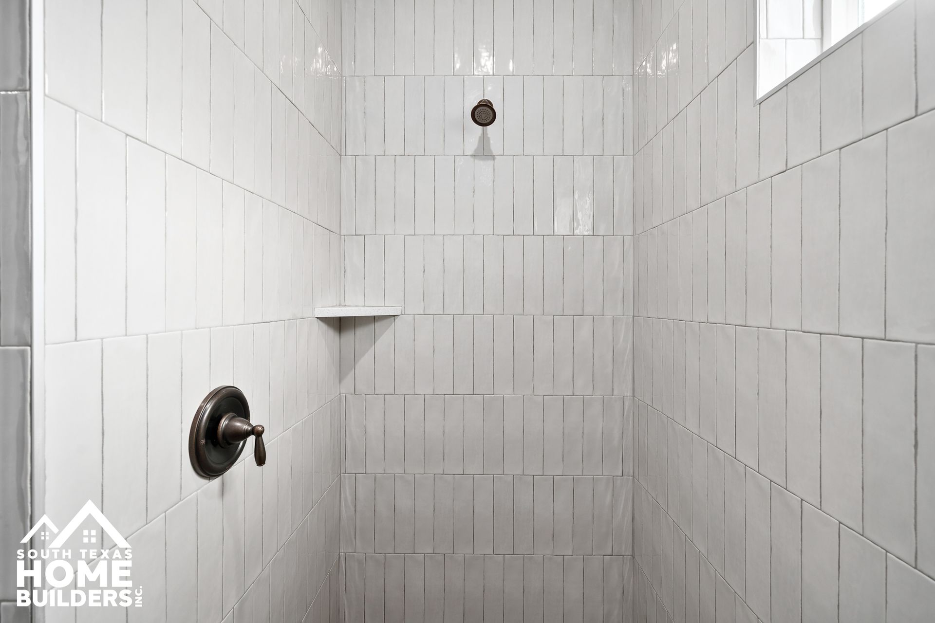 White-tiled shower with a dark faucet, showerhead, and a small shelf.