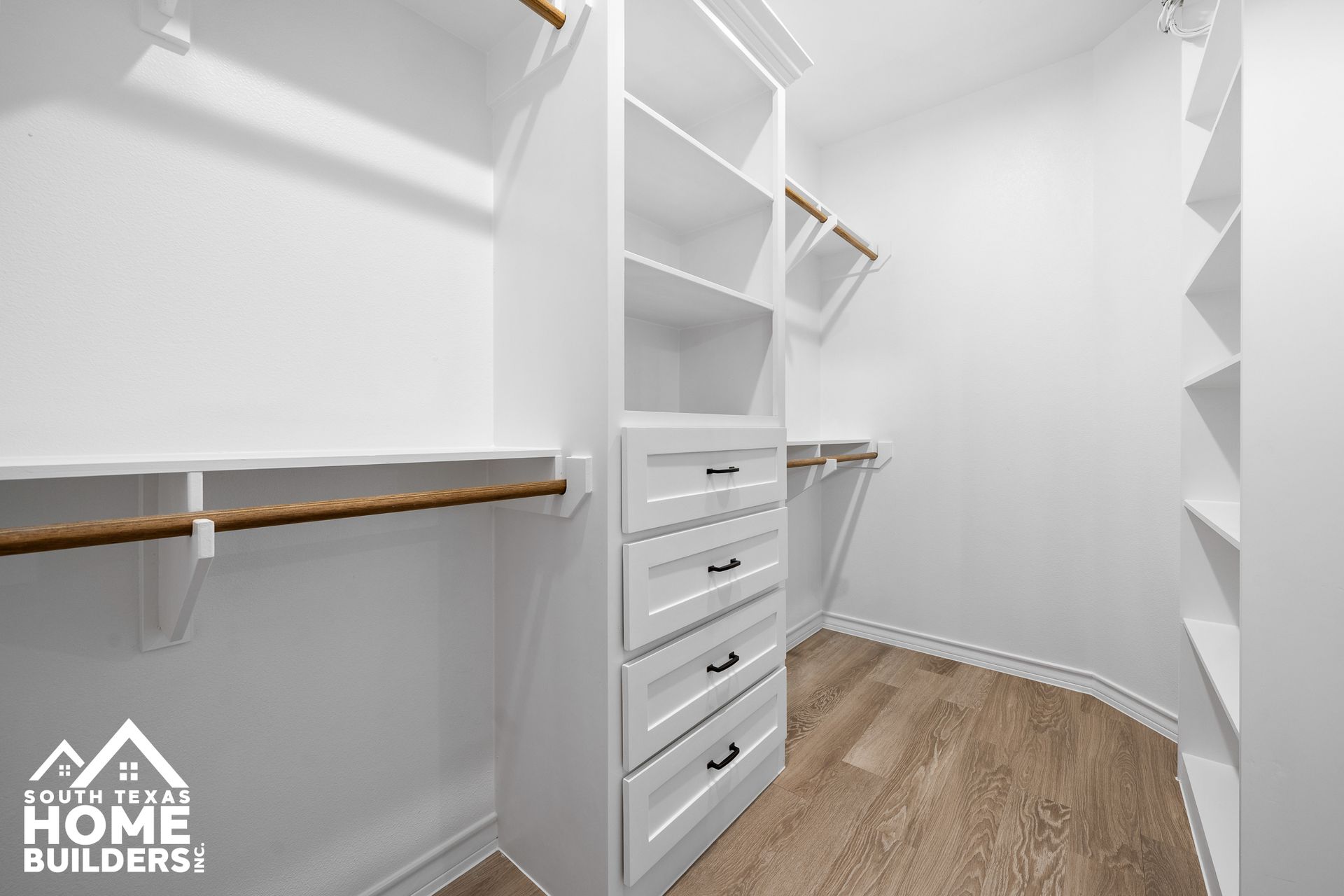 Walk-in closet with white shelves, drawers, and wooden rods. Light wood floor.