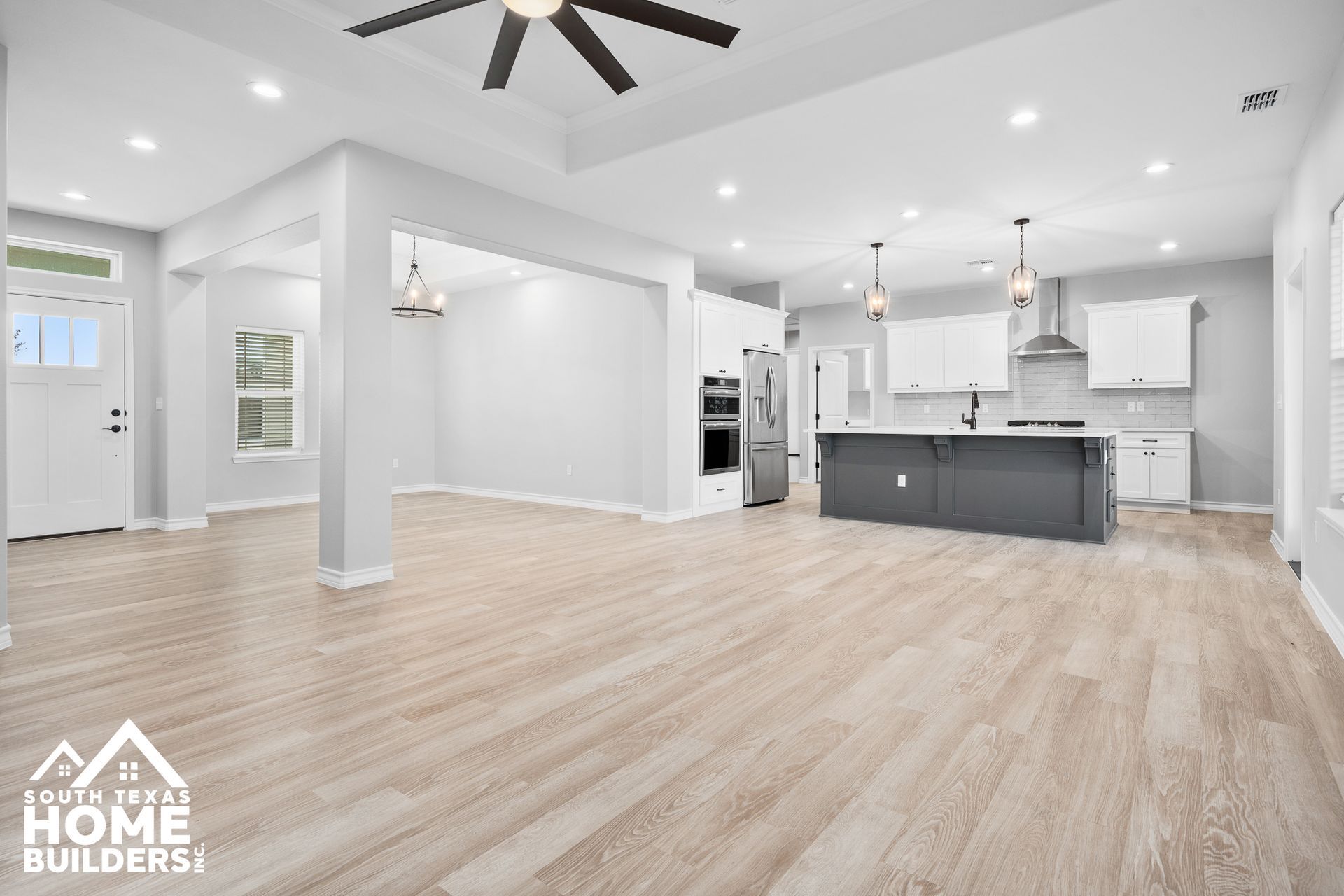 Spacious open-concept living area with light wood floors, kitchen, and white walls.