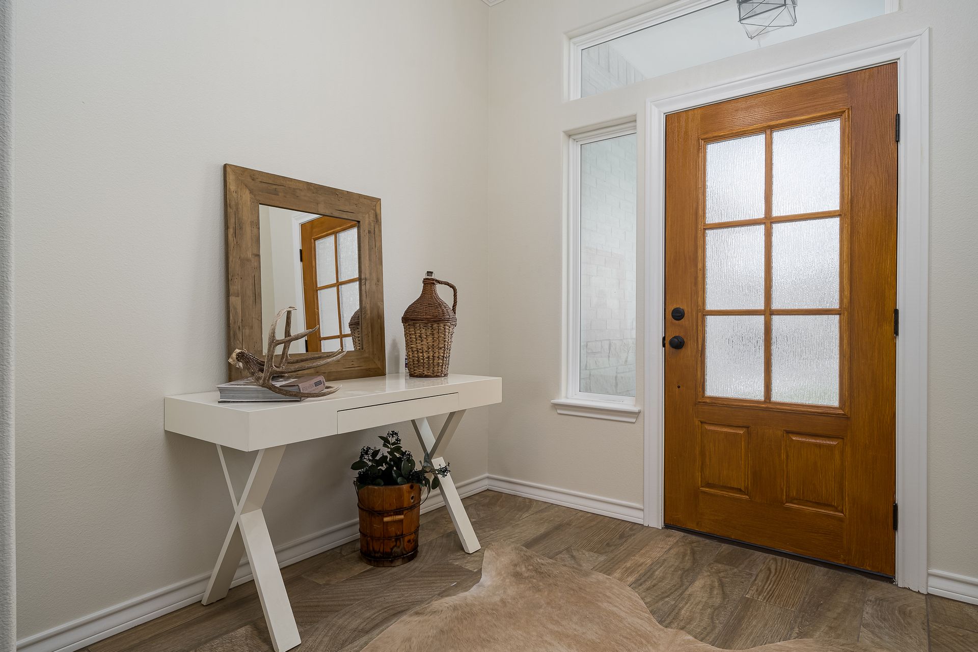 A hallway with a wooden door , a table , a mirror and a window.