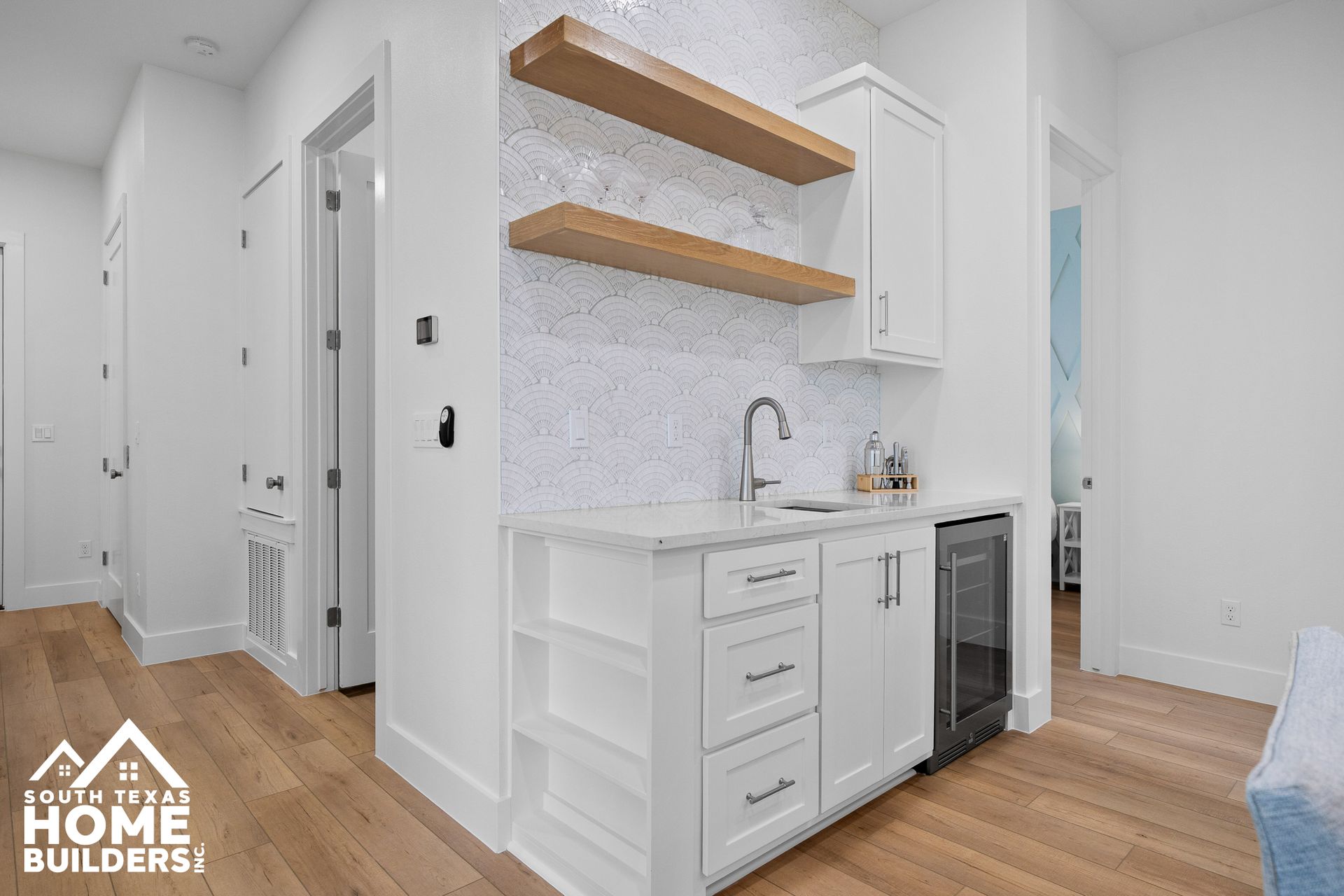 White home bar with shelves, sink, and mini-fridge in a hallway.