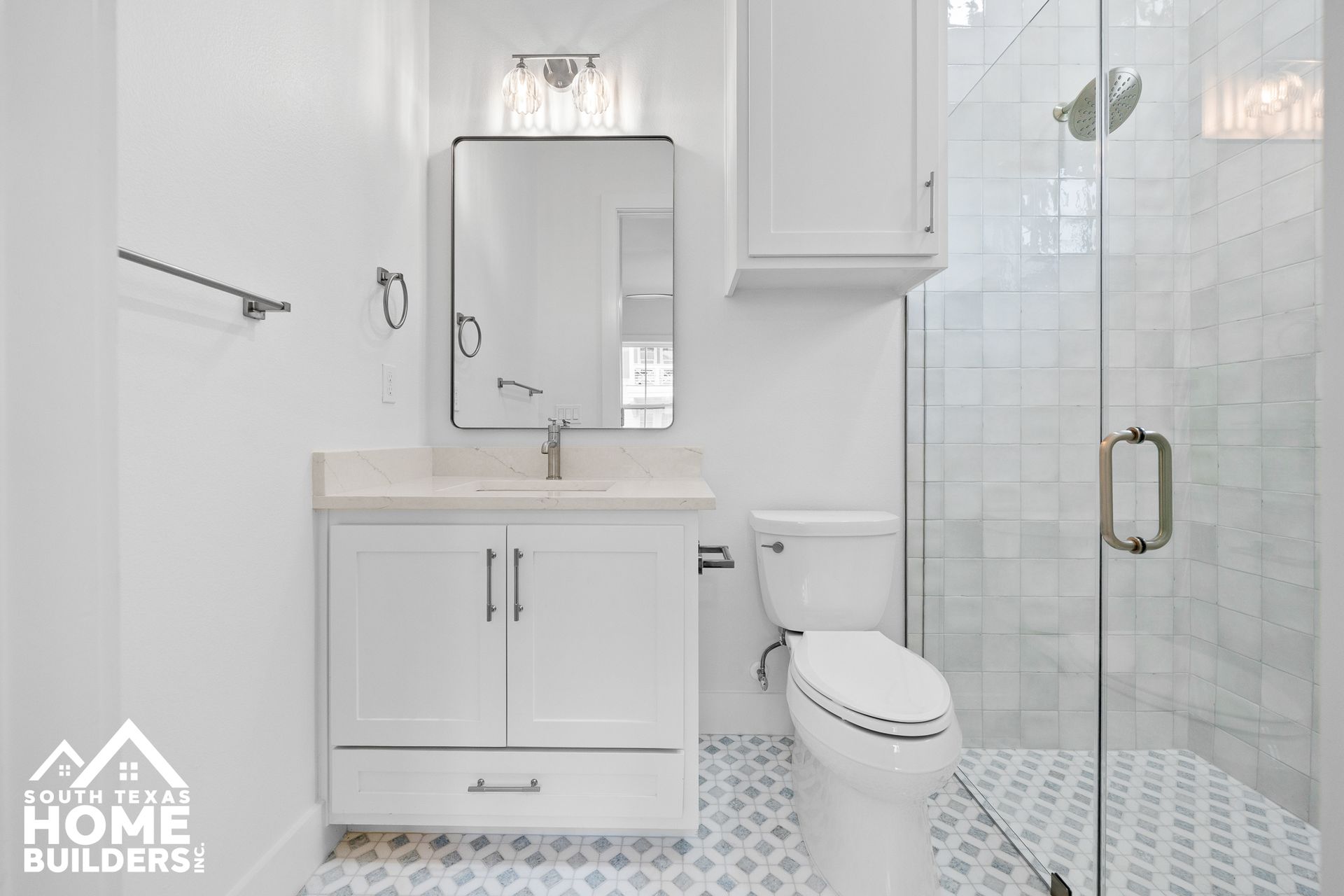 White bathroom with vanity, toilet, and glass shower. Blue and white patterned floor tiles.