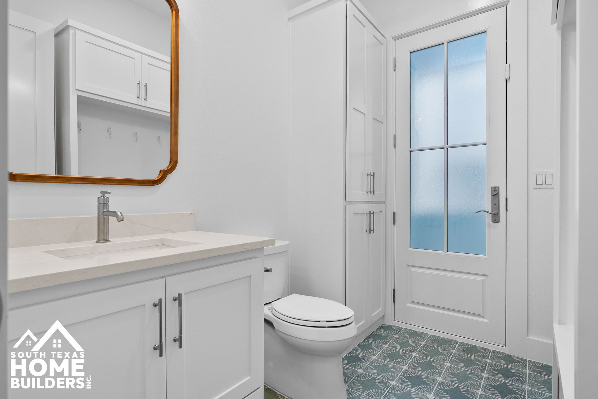 White bathroom with vanity, toilet, and storage; gold-framed mirror; teal patterned floor; door with frosted glass.