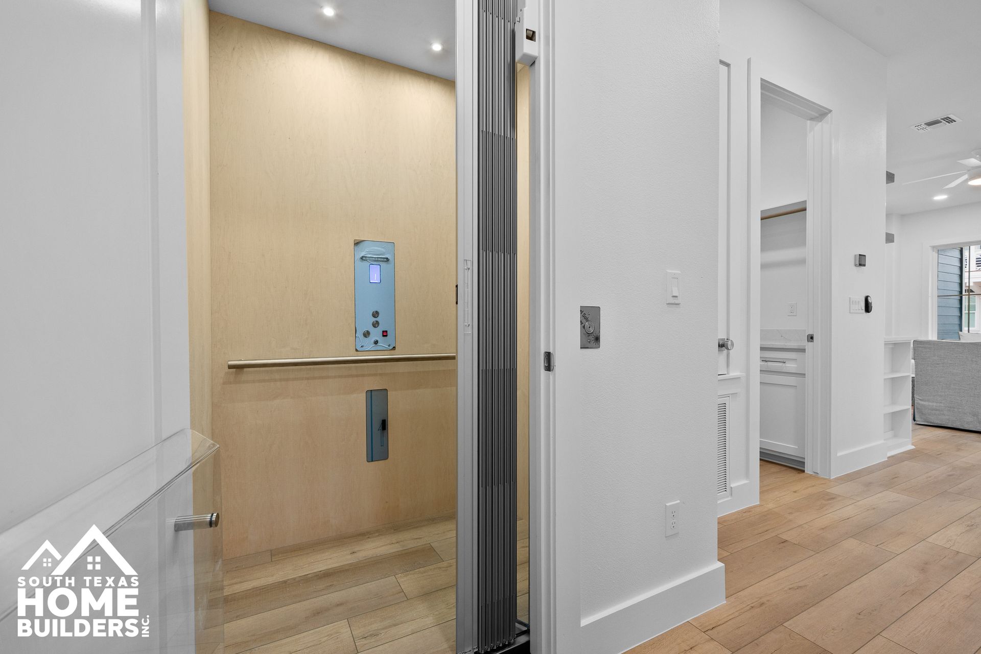 Interior view of an elevator with a light-colored wooden interior, next to a white hallway with wooden flooring.