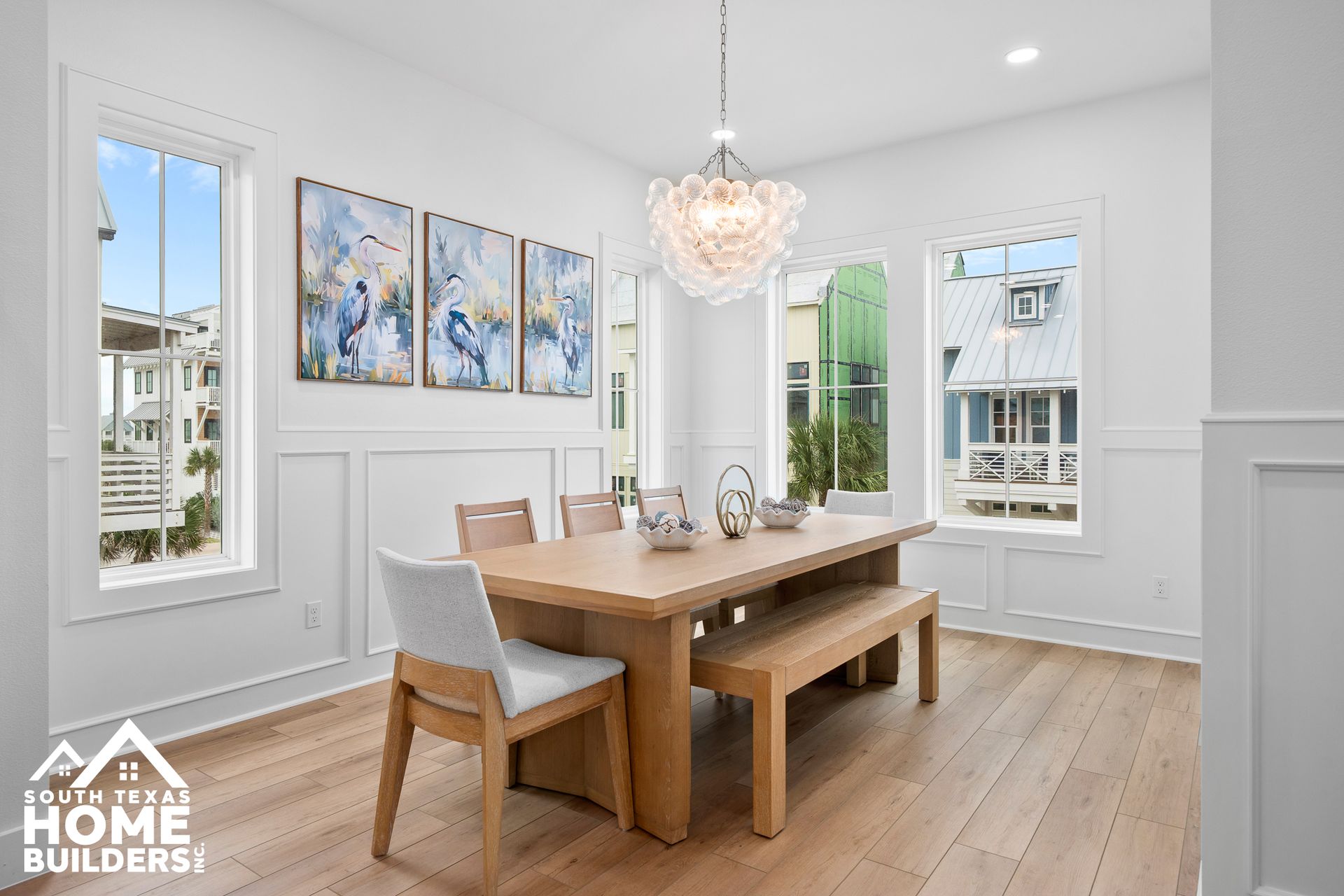 Dining room with wooden table, bench, chairs, art, and chandelier. White walls, wood floor, and windows.