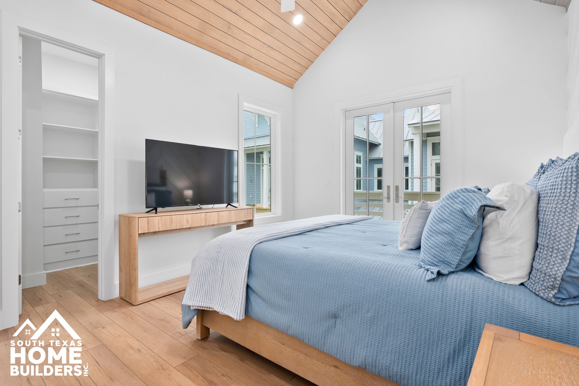 Bedroom with a blue bed, TV, light wood floors, and vaulted wood ceiling.