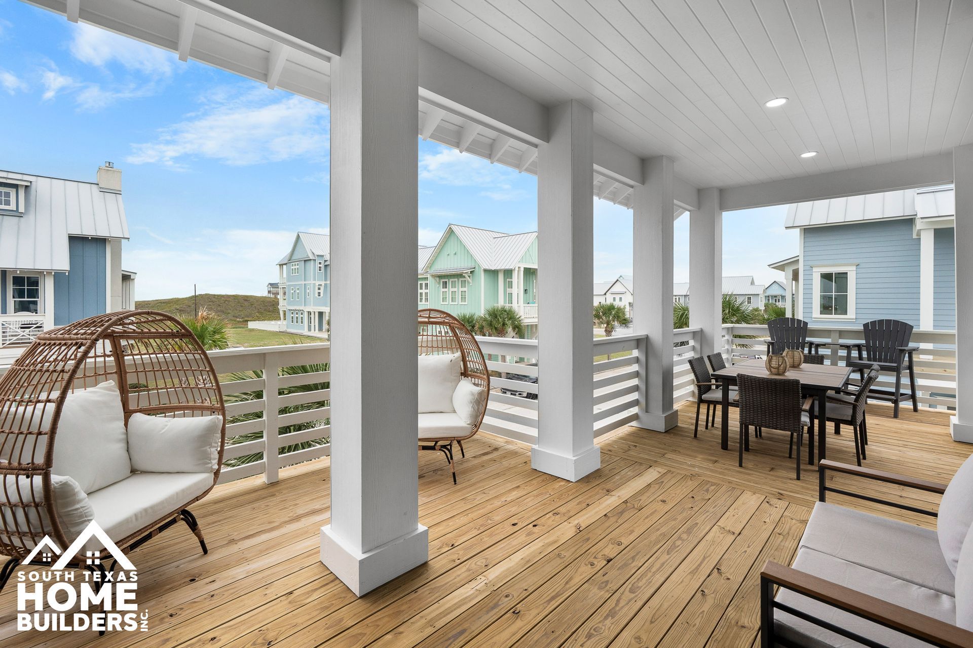 Spacious porch with seating, overlooking houses and a grassy area, under a blue sky.