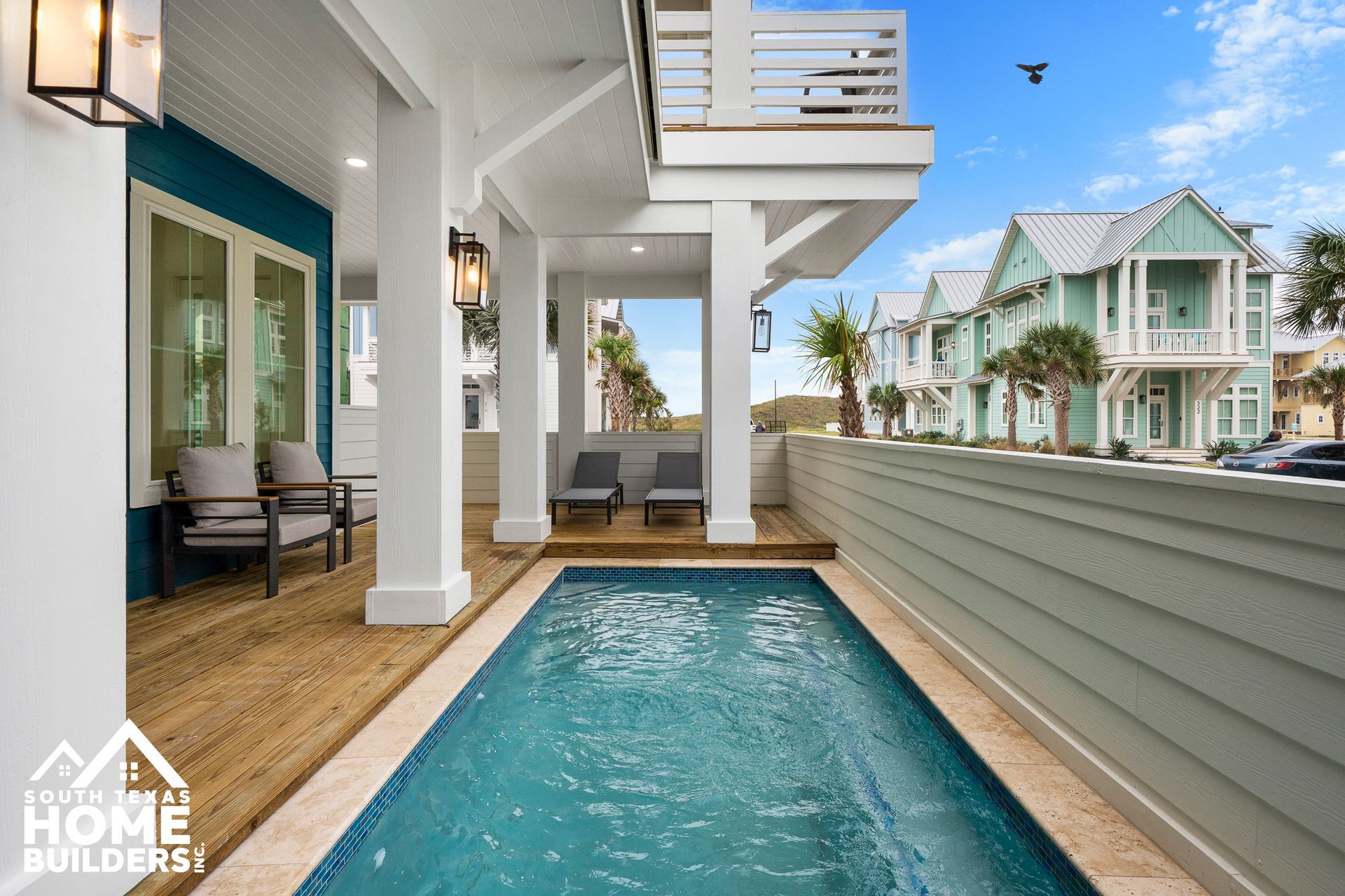 A narrow swimming pool under a covered patio with teal siding and multiple beach homes in the background.