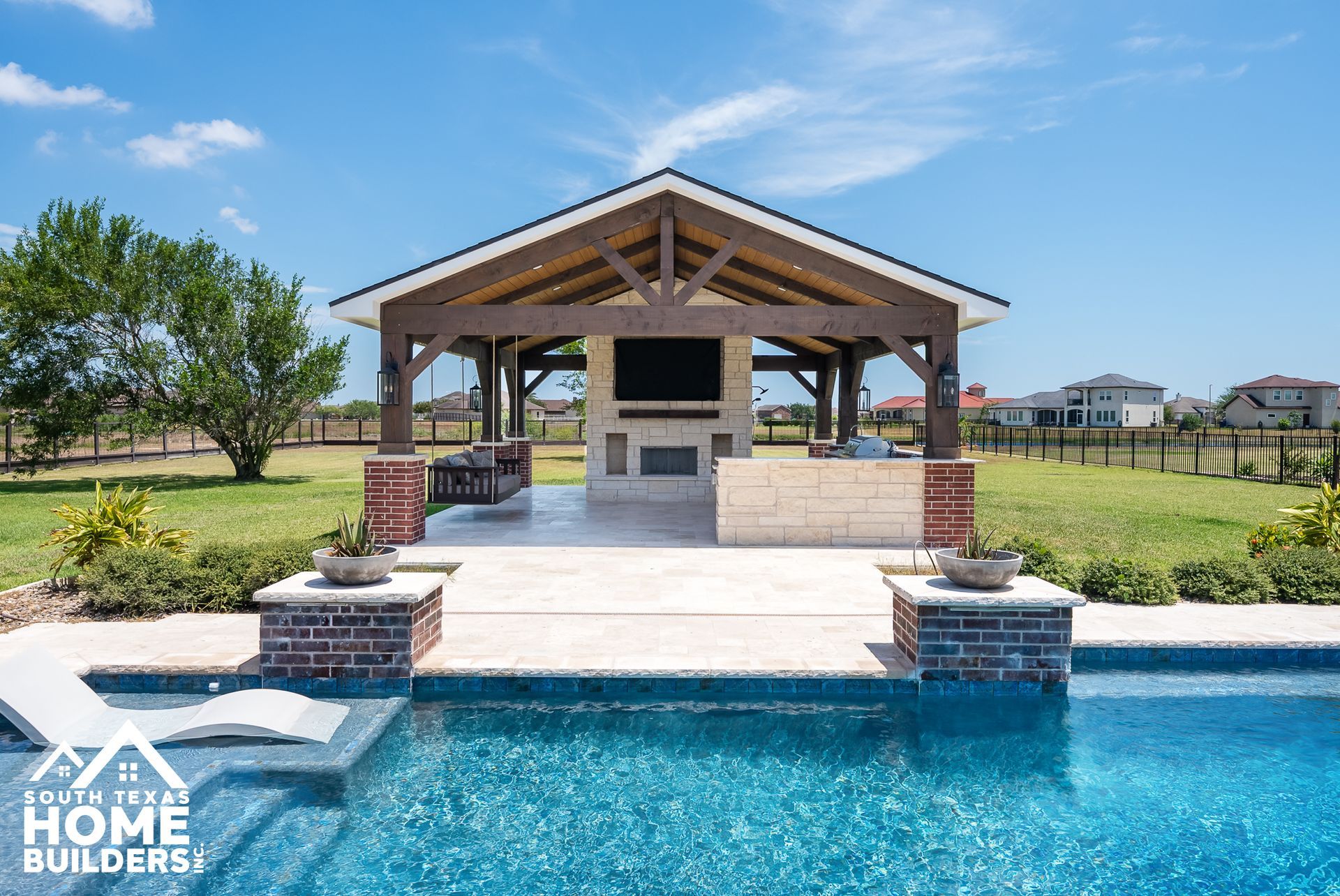 Outdoor patio with a pool, fireplace, and seating under a wooden pergola on a sunny day.