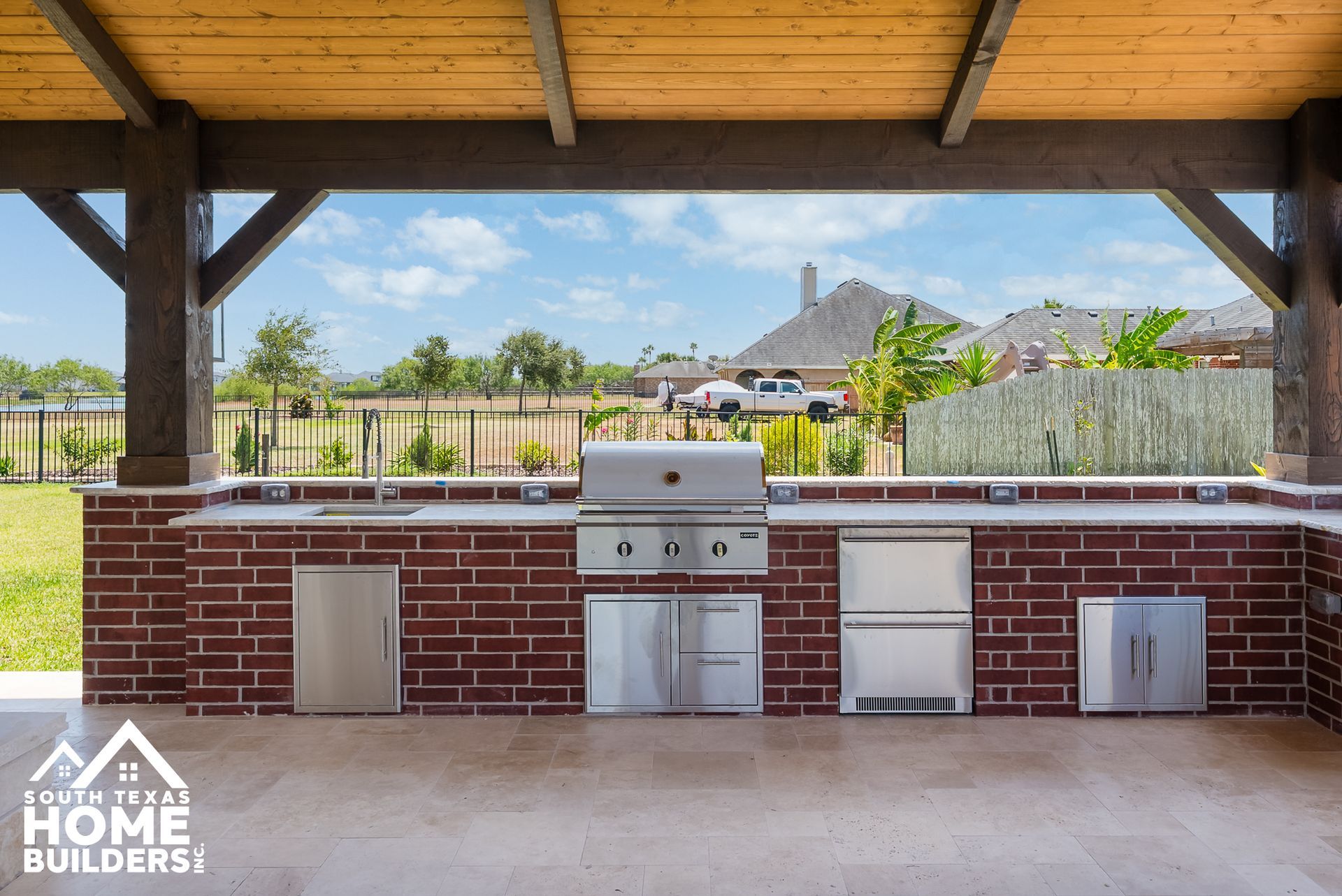 Outdoor kitchen with brick facade, grill, sink, and fridge under wooden pergola, overlooking a backyard.