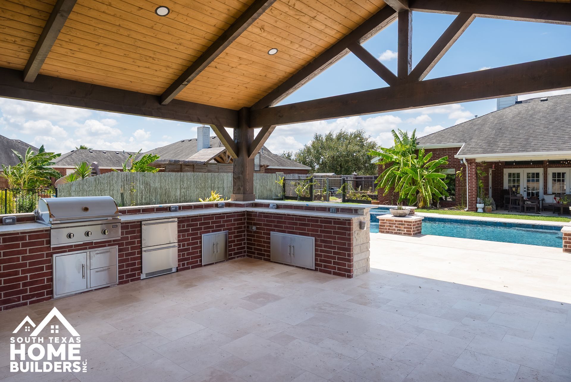 Outdoor kitchen with brick facade, stainless steel appliances, and a wooden overhead structure.