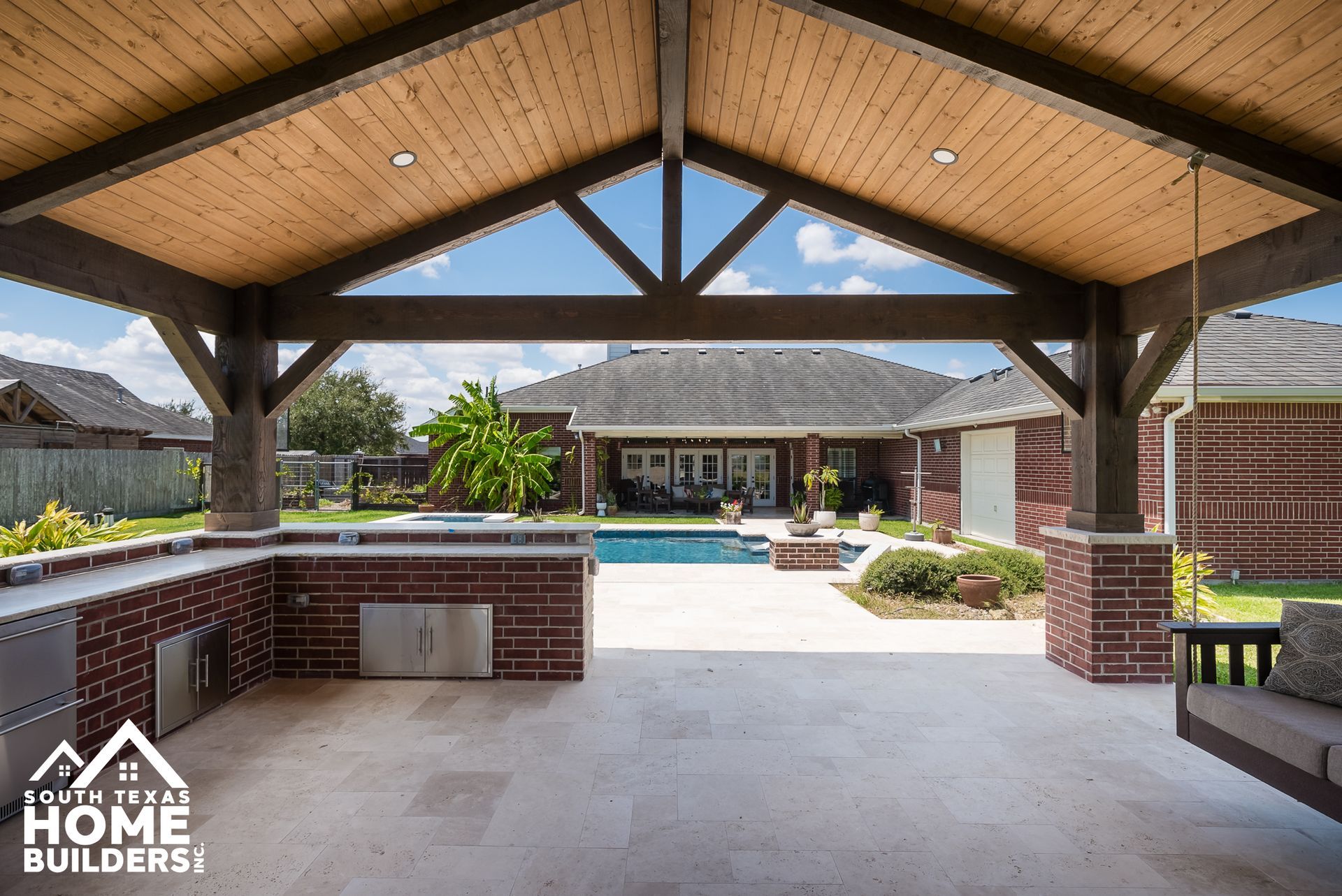 Outdoor kitchen and patio with brick facade, pool, and pergola with wooden beams.