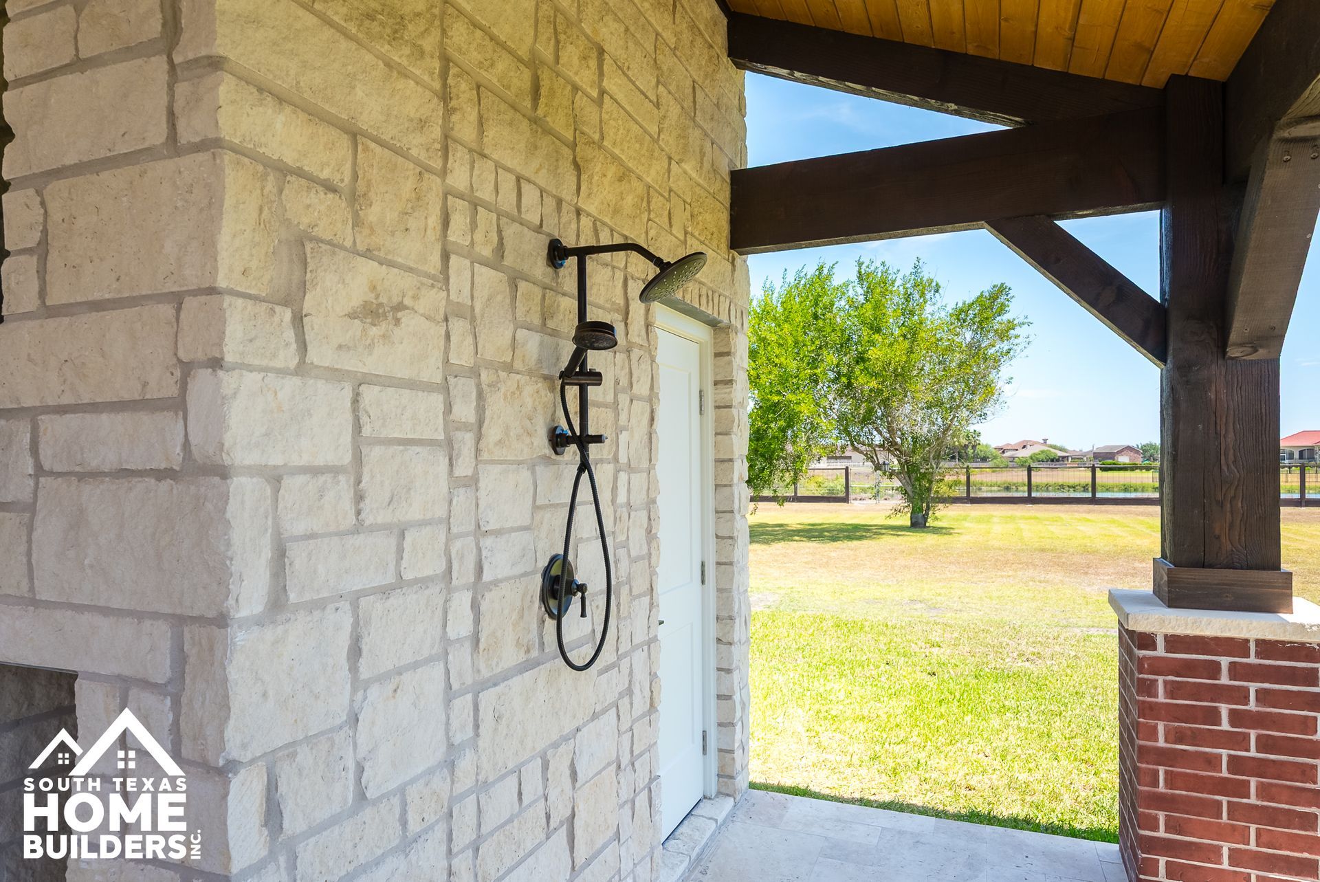 Outdoor shower on a stone wall, near a grassy yard and wooden porch.
