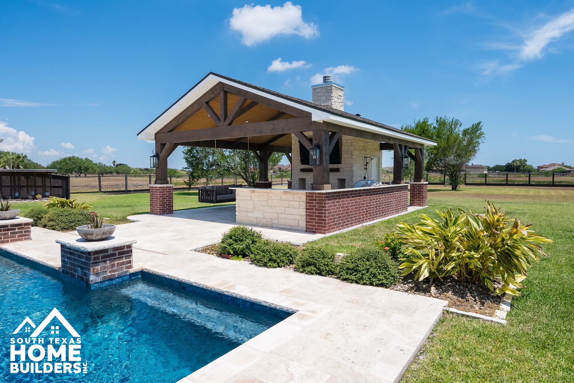 Outdoor kitchen and pool area with a covered structure, brick, and stone features under a sunny sky.