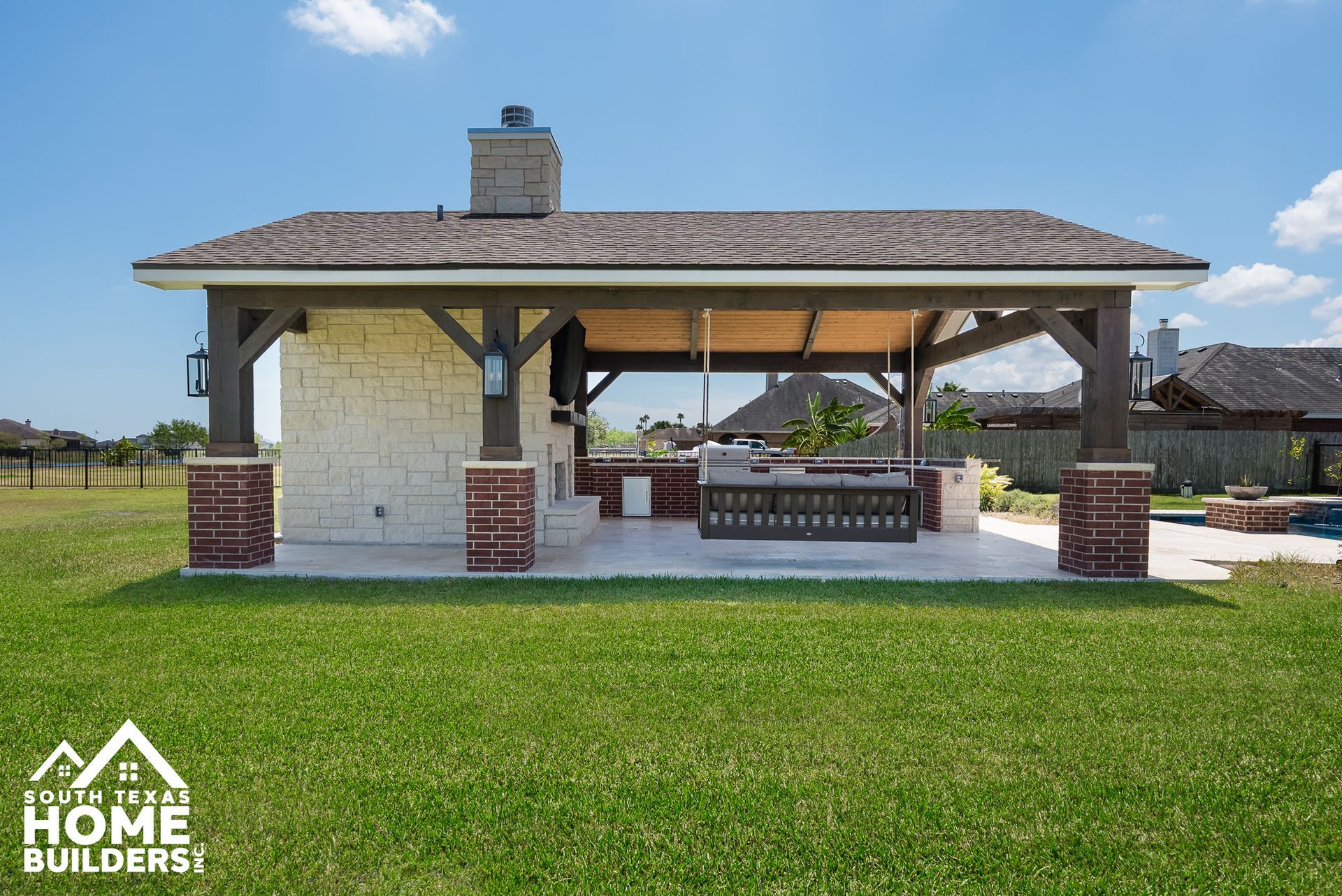 Outdoor kitchen under a dark wood-framed roof with stone and brick columns and a green lawn.