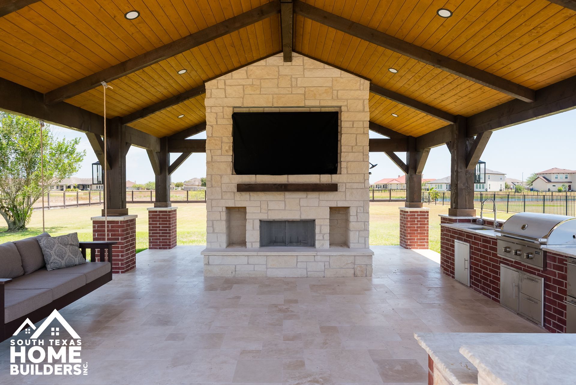 Covered outdoor kitchen with fireplace, TV, seating, and grill; light stone, wood beams.