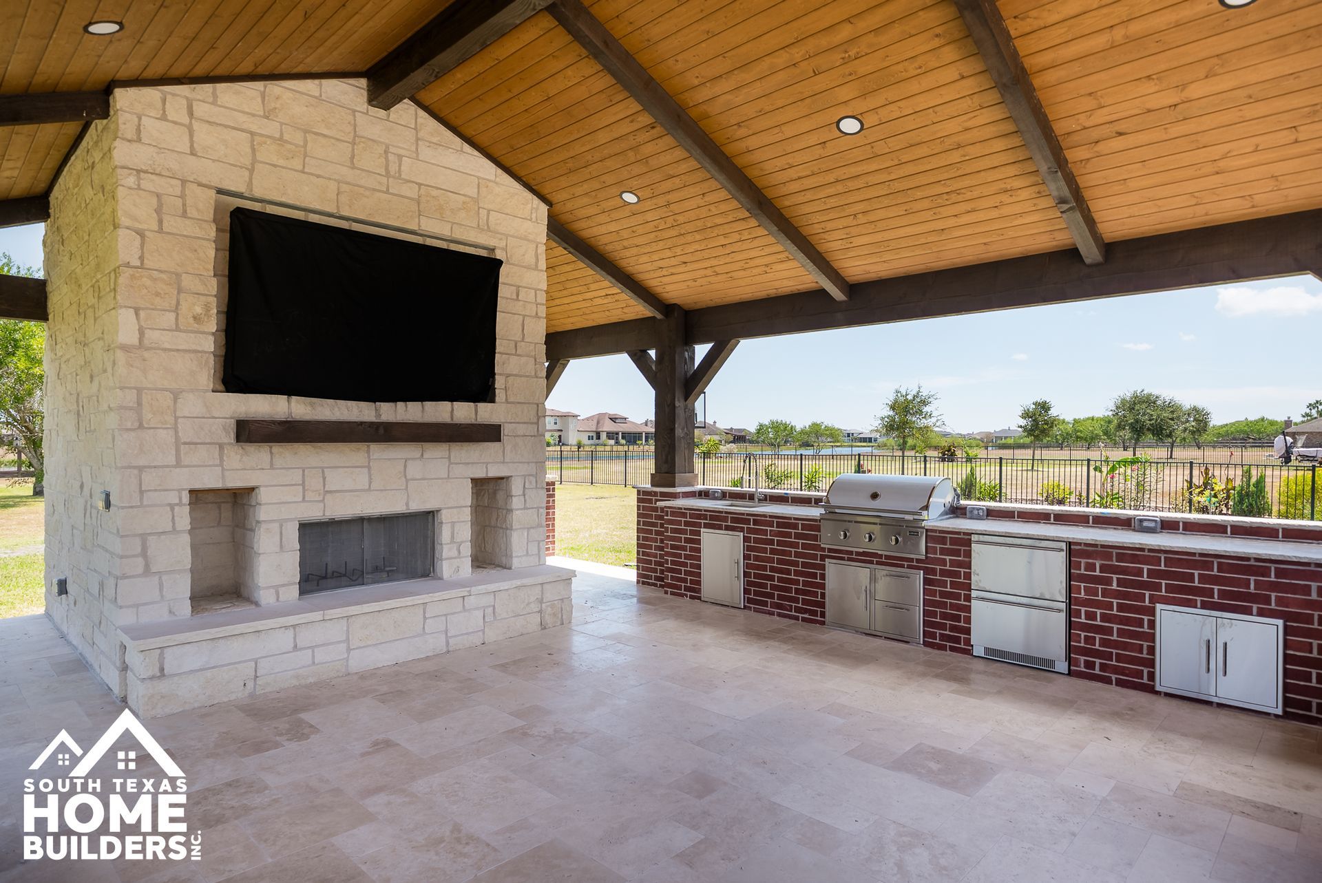 Outdoor kitchen and fireplace with a large TV, brickwork, and stone facade, in a covered patio area.