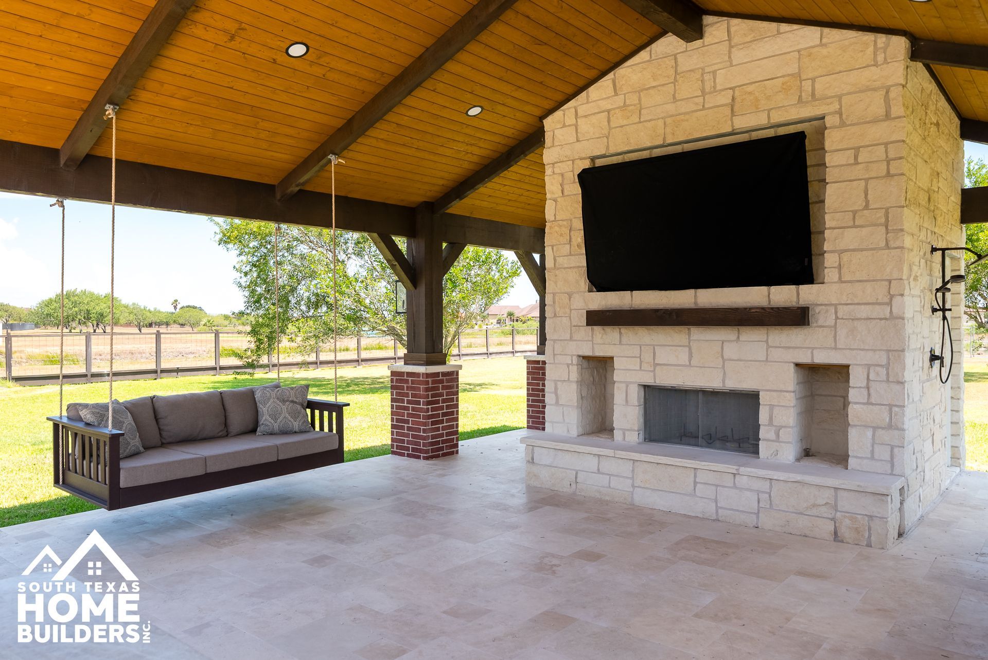 Outdoor patio with stone fireplace, TV, and wooden swing. Beige stone, brown wood, and a green lawn.