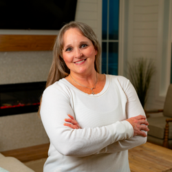 A woman in a white sweater is standing in a living room with her arms crossed.