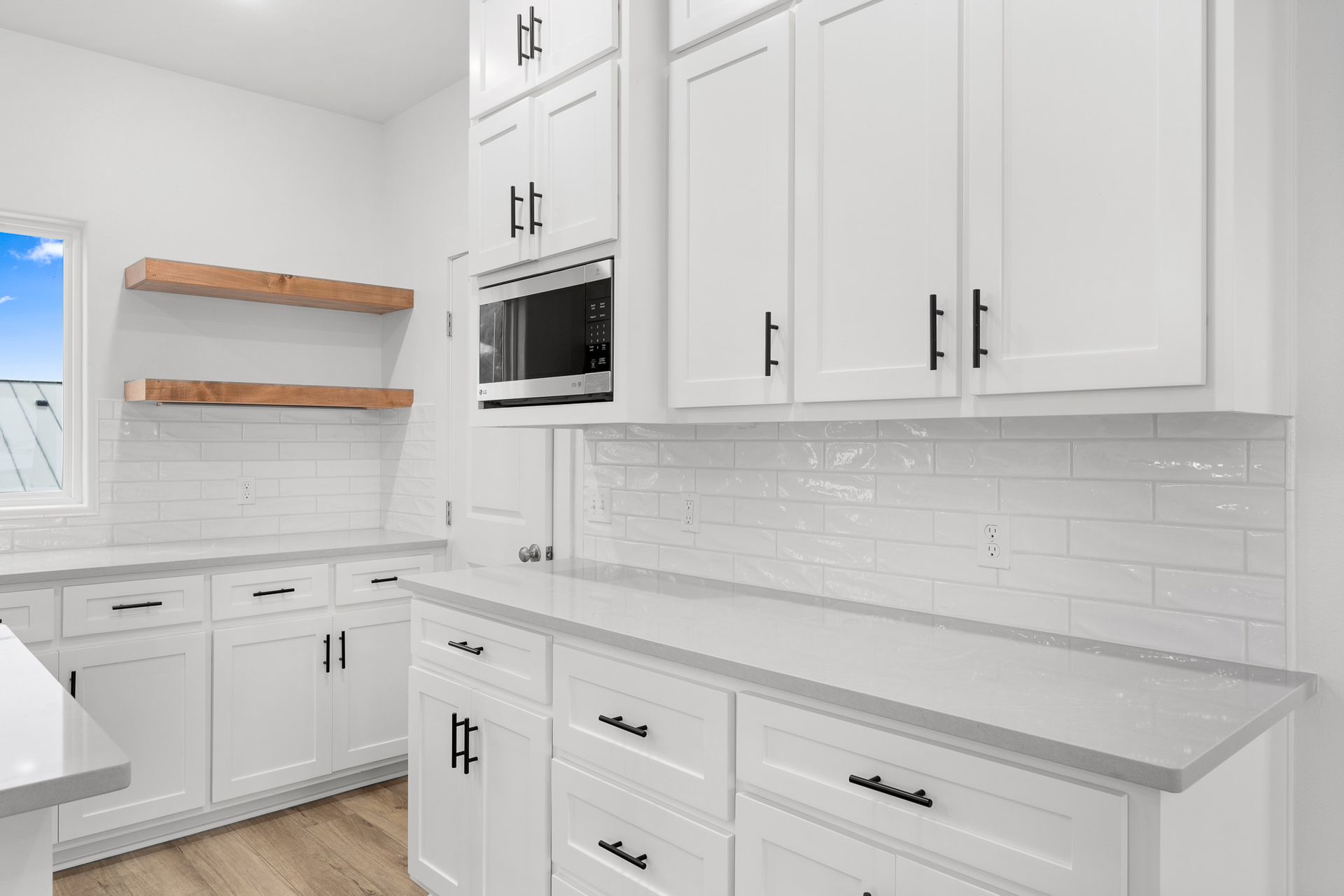 A kitchen with white cabinets , white counter tops , a microwave and a window.