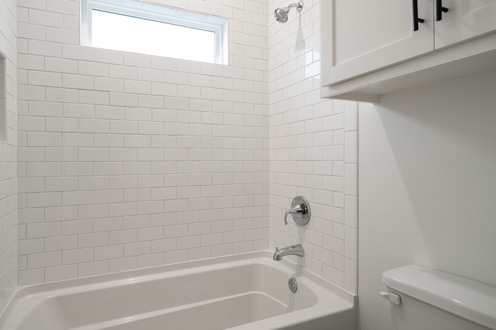 A bathroom with white tiles , a tub , a toilet and a window.