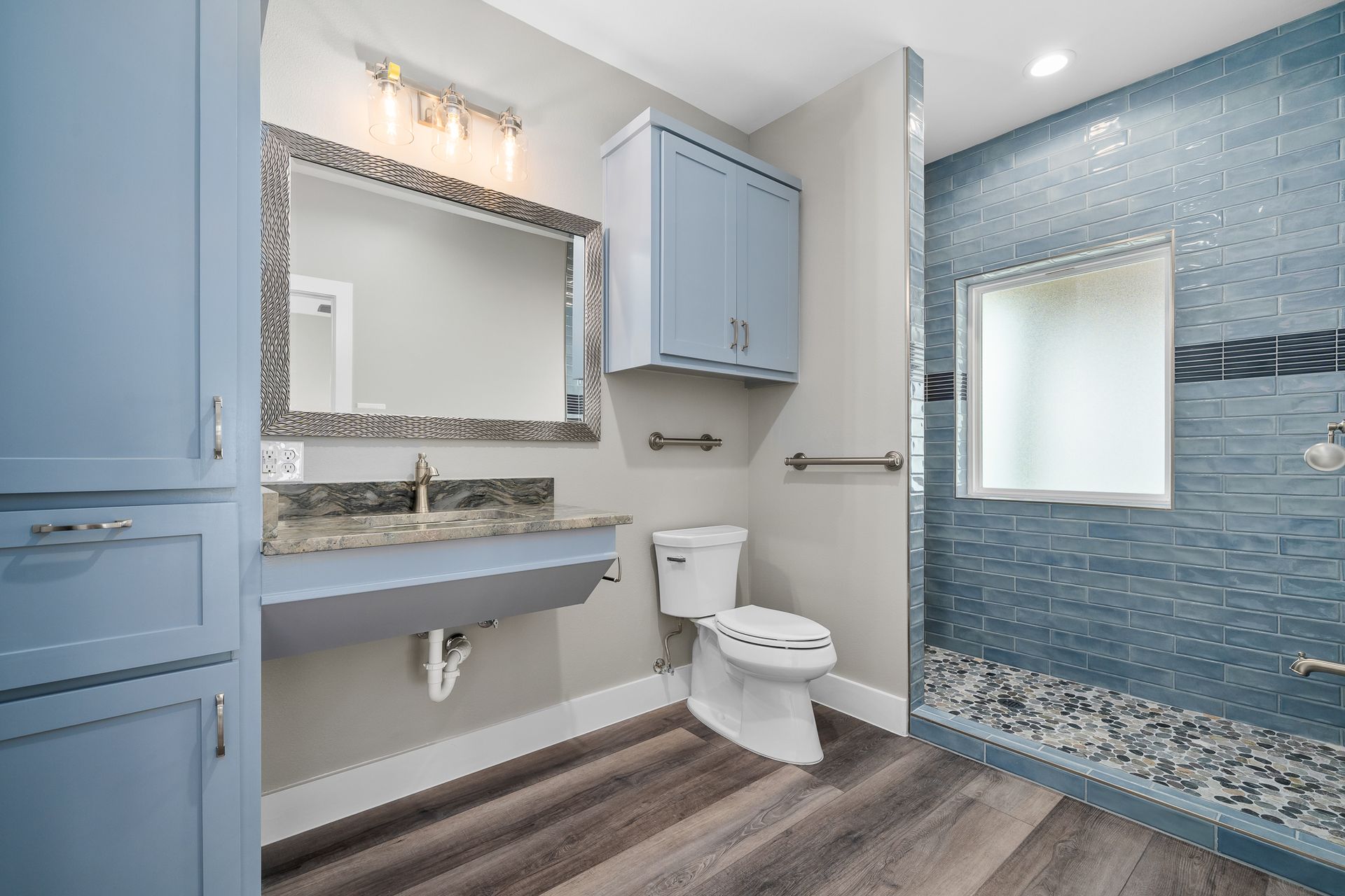 Accessible bathroom with blue cabinets, sink, toilet, and tiled shower with a window.
