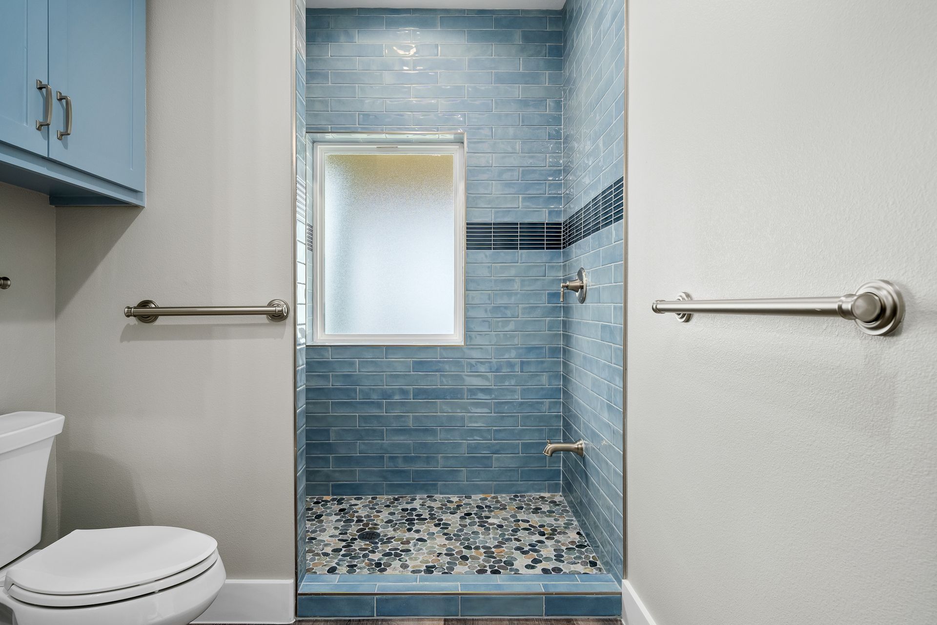 Bathroom with blue tiled shower, pebble floor, and frosted window. Light blue cabinets and white toilet.