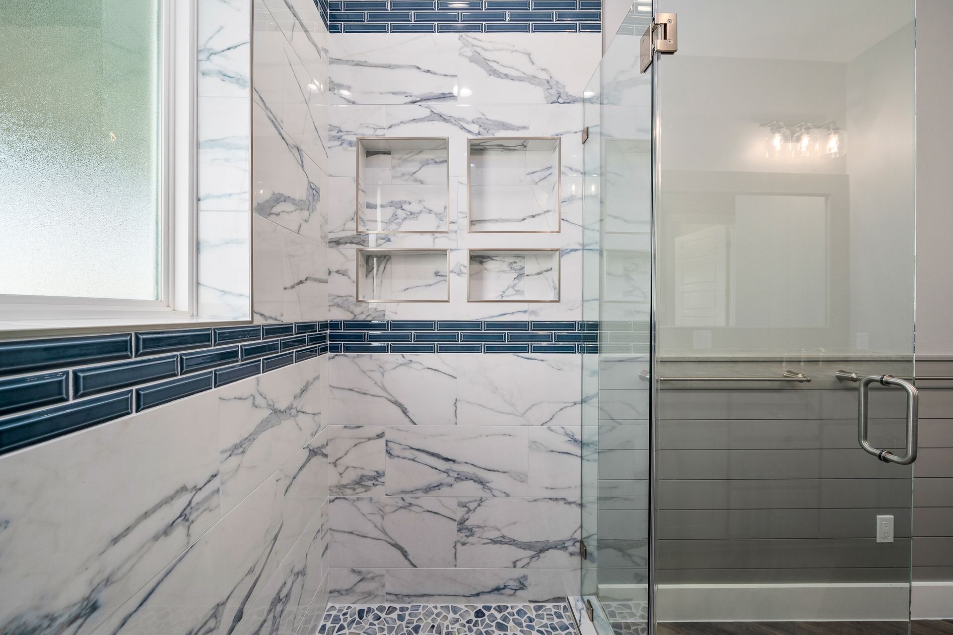 Shower with marble-look tile, blue accent tile, and glass door; built-in shelves and a window visible.