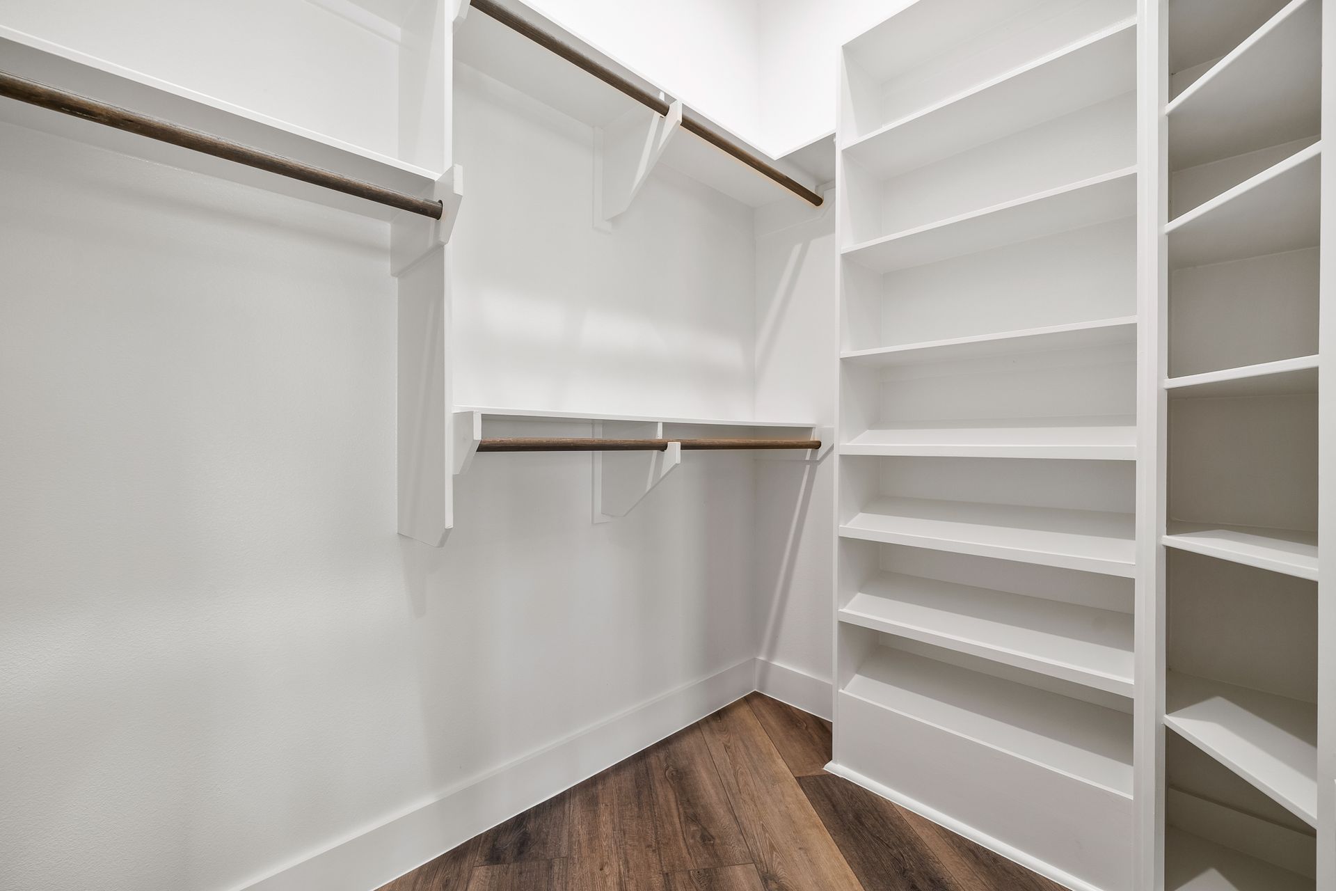 Empty white walk-in closet with shelves, hanging rods, and wood-look flooring.