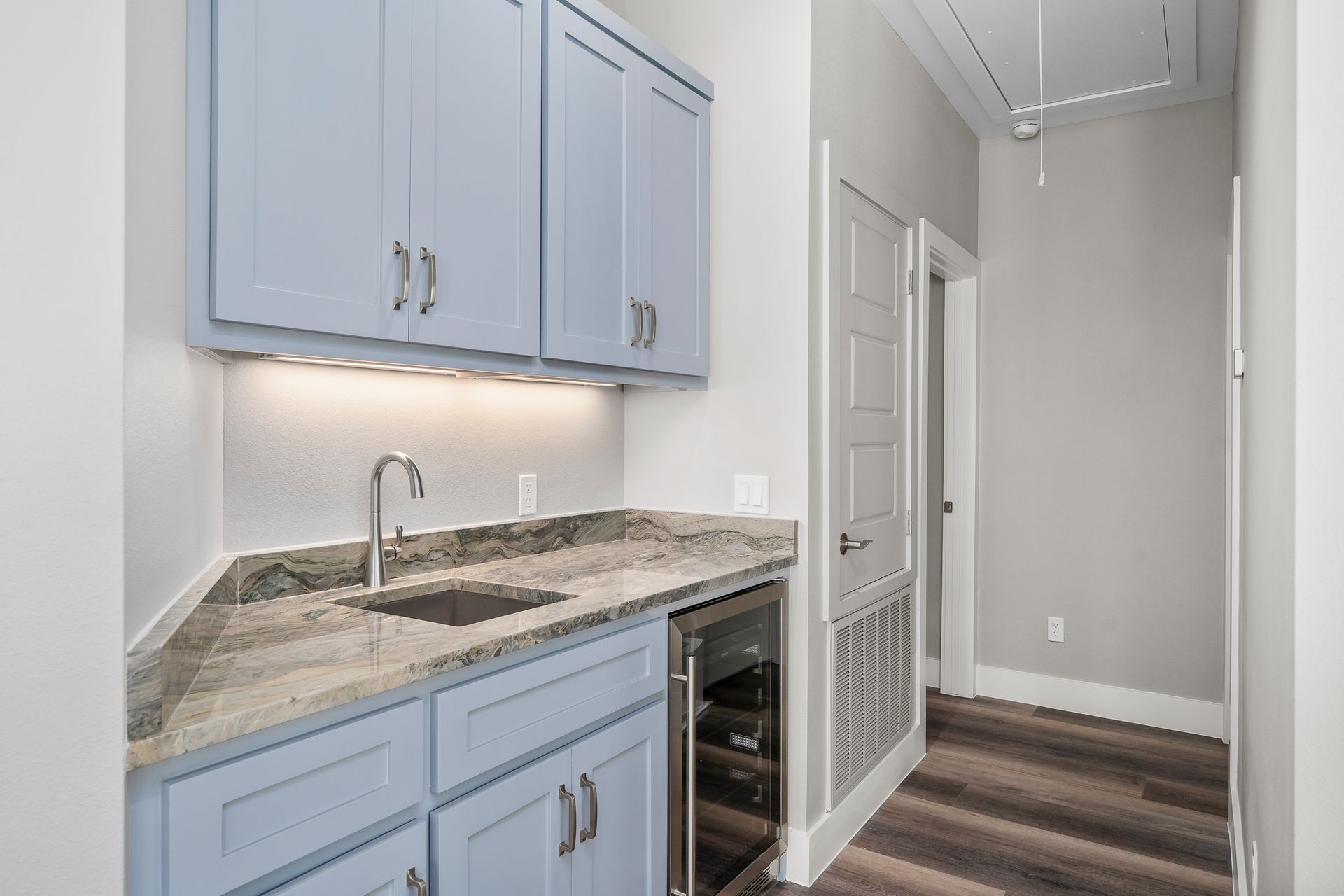 Light blue cabinets with a granite countertop and sink. A small refrigerator, hallway, and door are also visible.