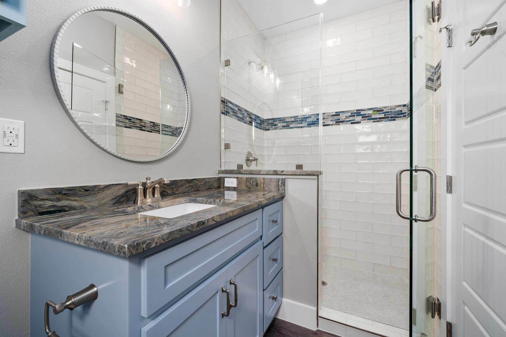 Bathroom with blue vanity, gray countertop, round mirror, glass shower with white tiles and blue accent.