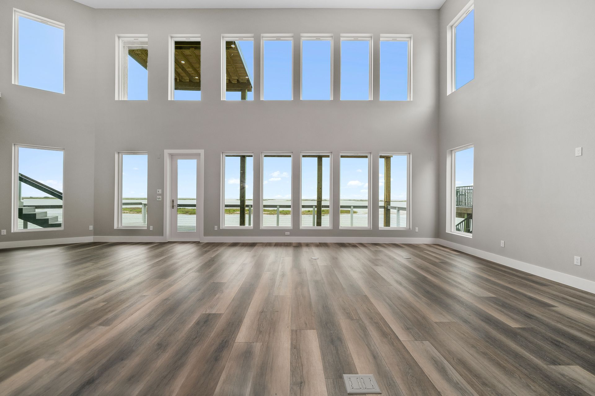 Empty living room with large windows overlooking water, gray walls, wood floors.