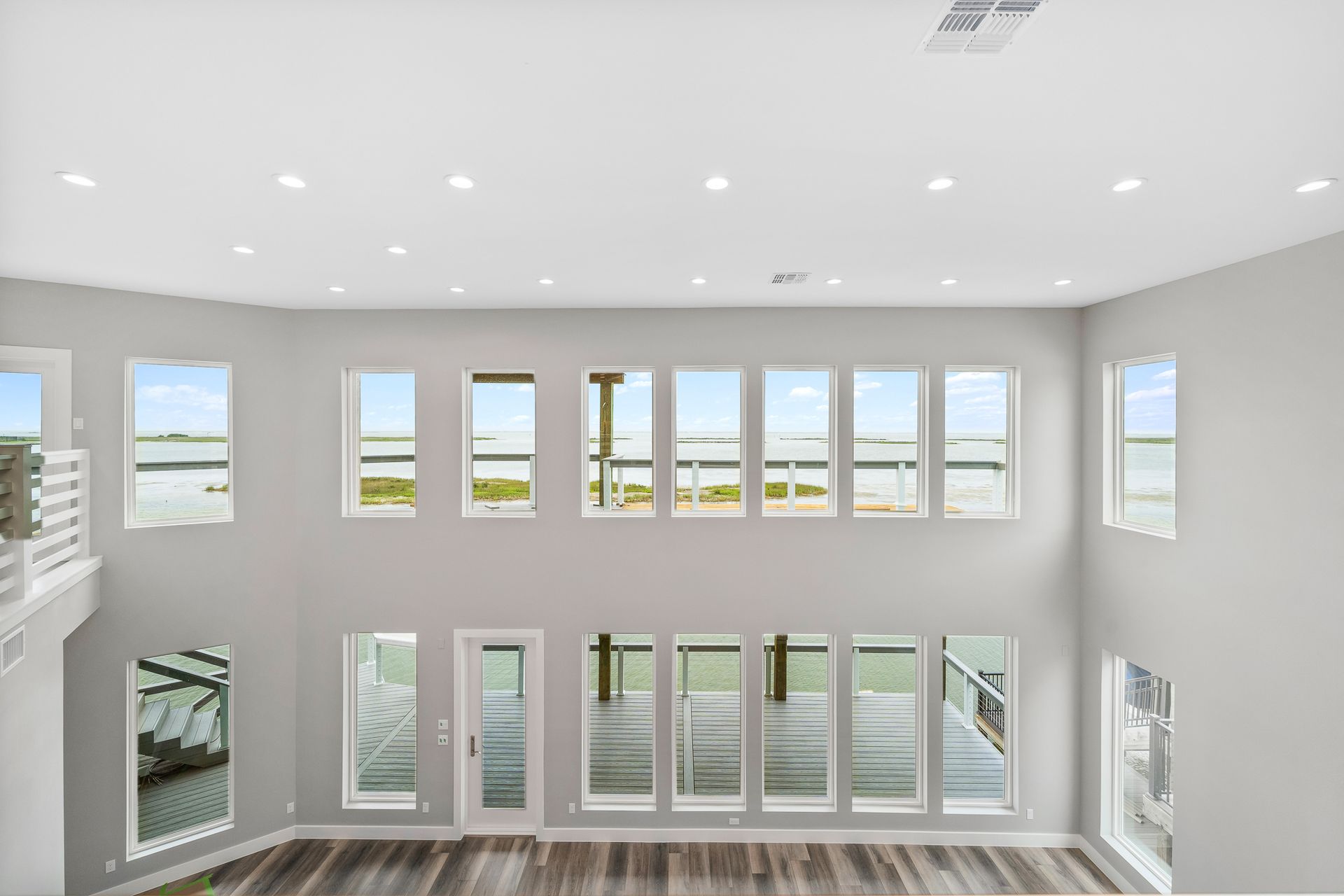 Spacious, light-filled living room with large windows overlooking water; gray walls, recessed lights, and wooden floor.