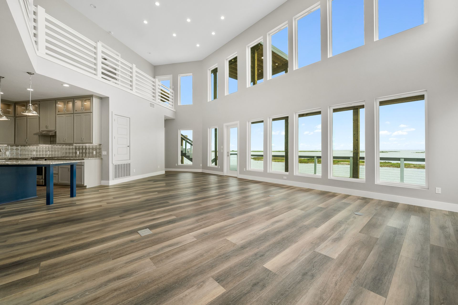 Open-concept living space with large windows overlooking a body of water, featuring wood flooring and a kitchen area.