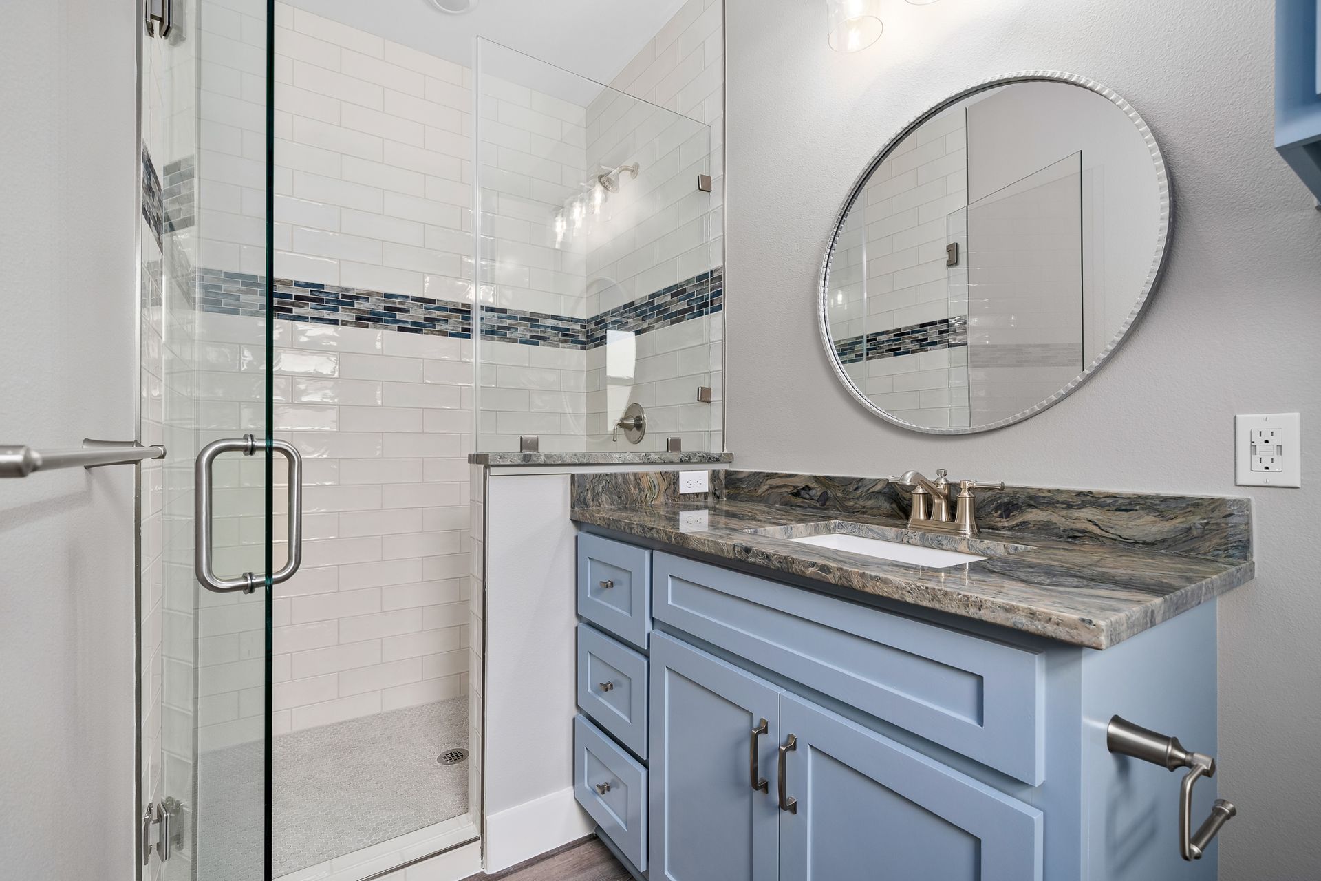 Bathroom with blue vanity, glass shower, granite countertop, and round mirror.