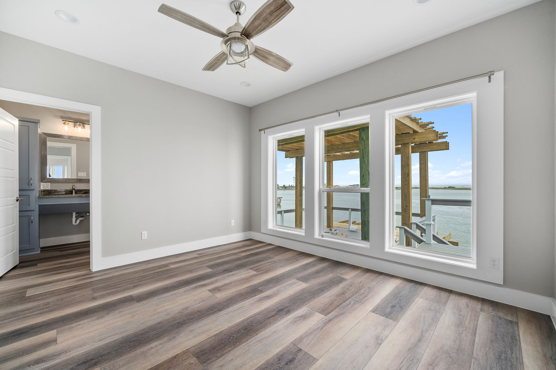 Bedroom with gray walls, wood floor, large windows overlooking water and dock; a door leads to a bathroom.