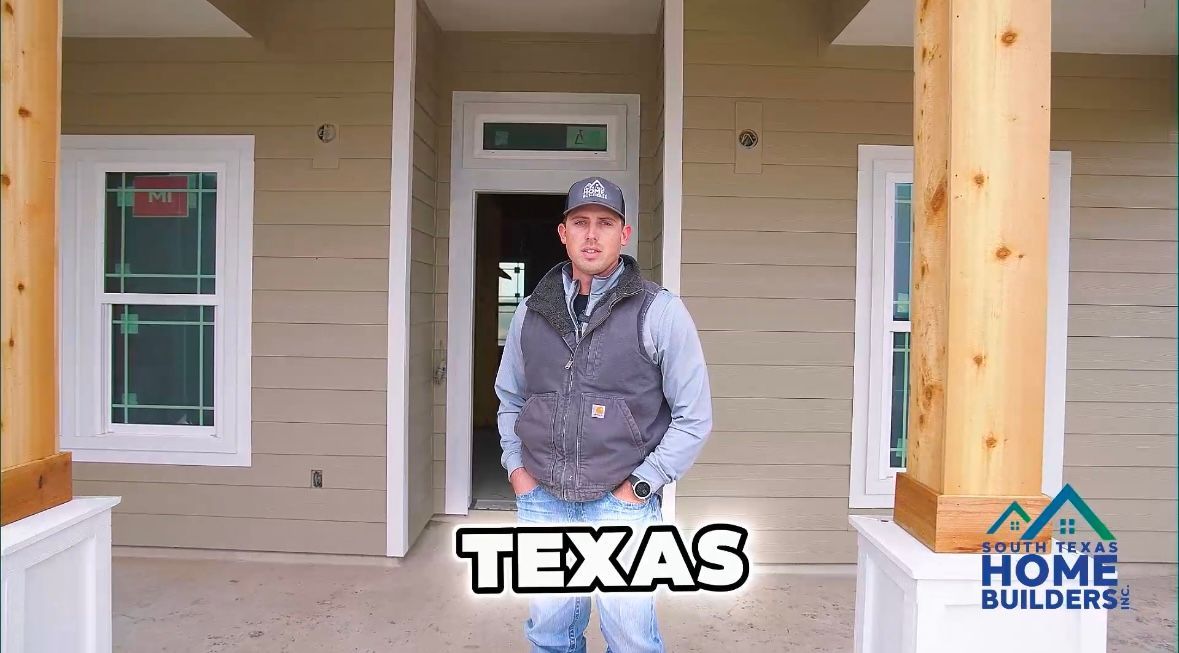 Man standing in front of a house in Texas. He's wearing a vest and jeans. House has wood columns, siding, and windows.