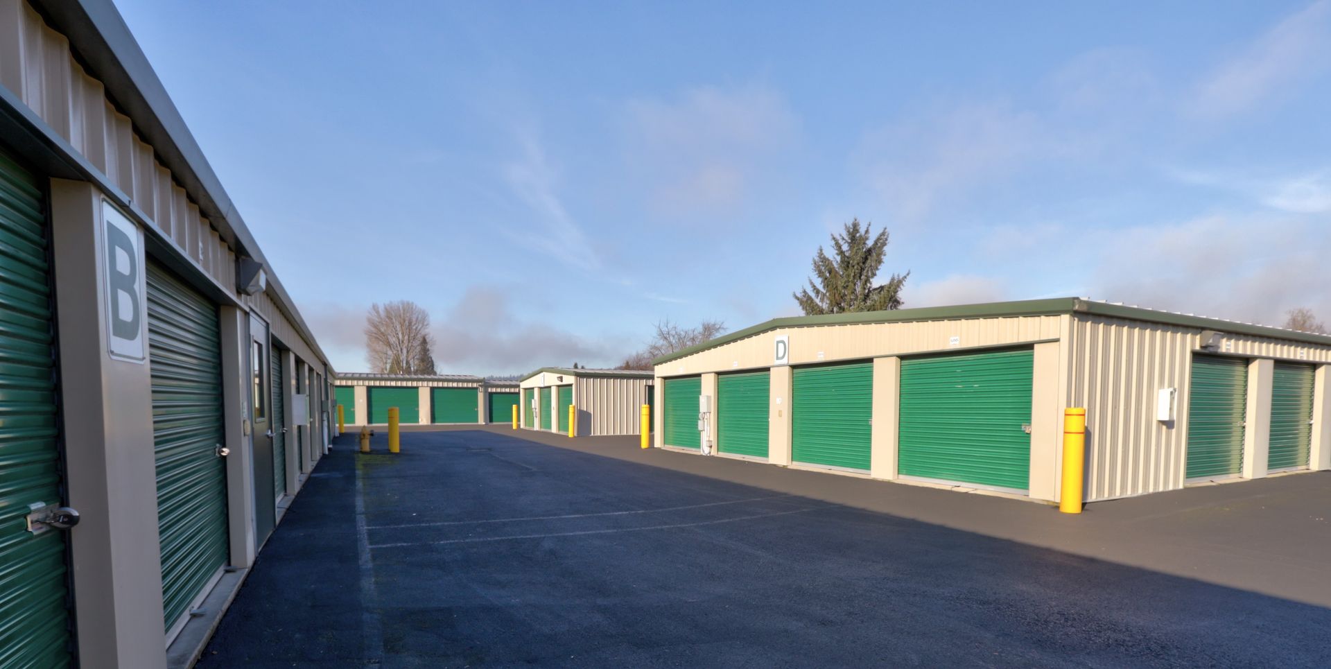 Exterior view of a storage facility with green doors and asphalt ground, under a cloudy blue sky.